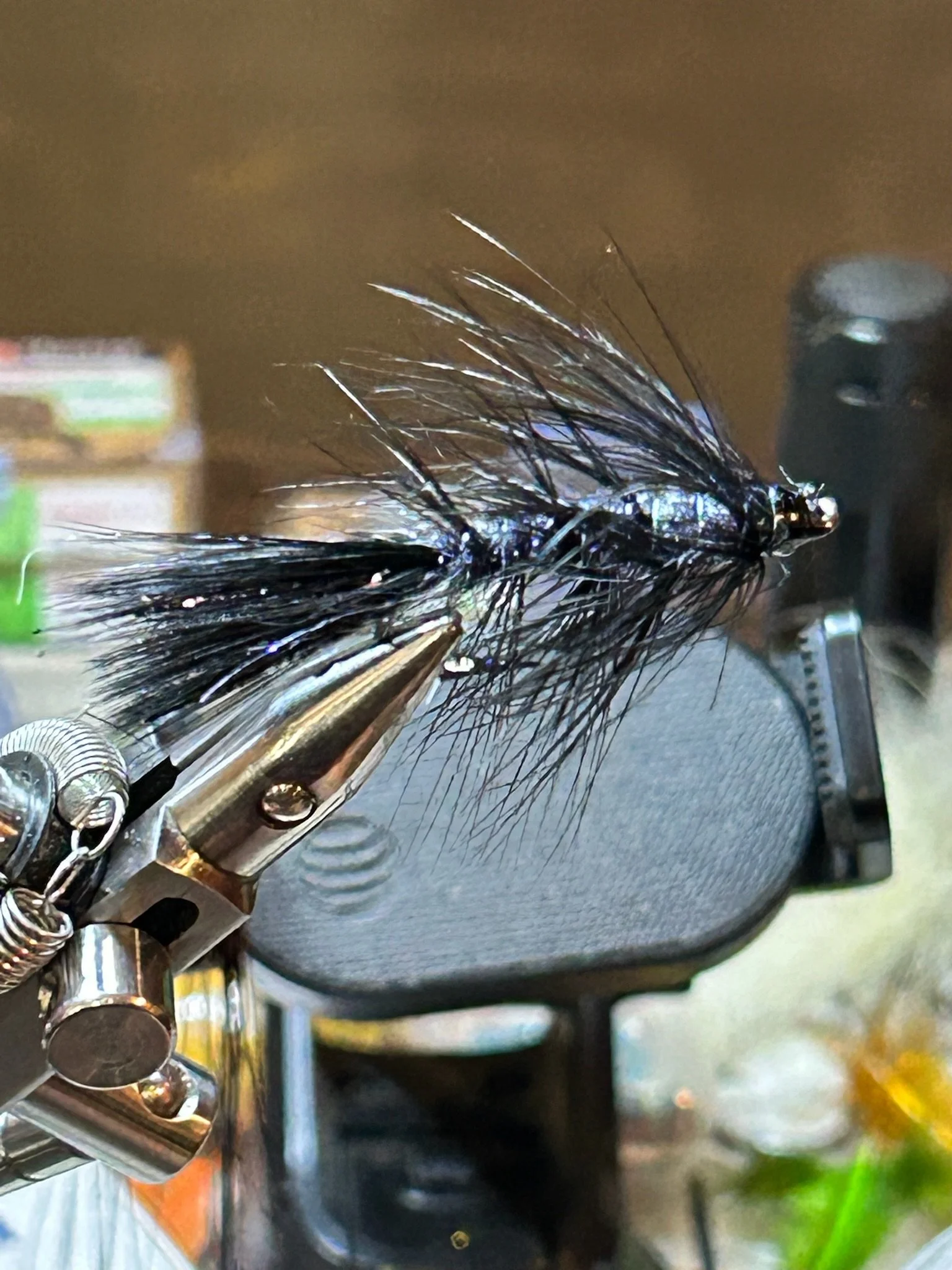 Close-up of a black fishing fly with shiny fibers, held in a fly-tying vise, with a blurred background.