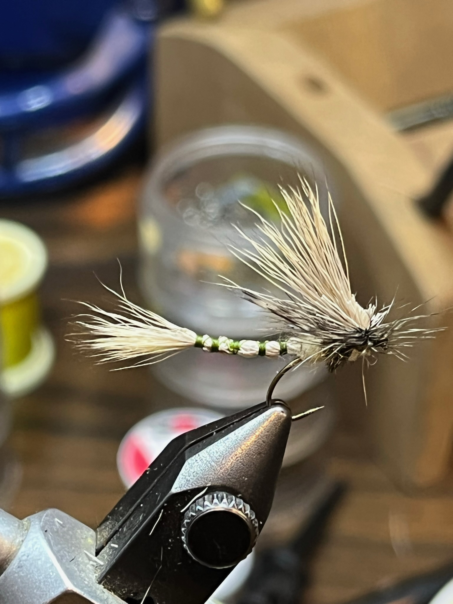 Close-up of a fishing fly held in a tool, with a blurred background showing a container and various objects, used for fly fishing.