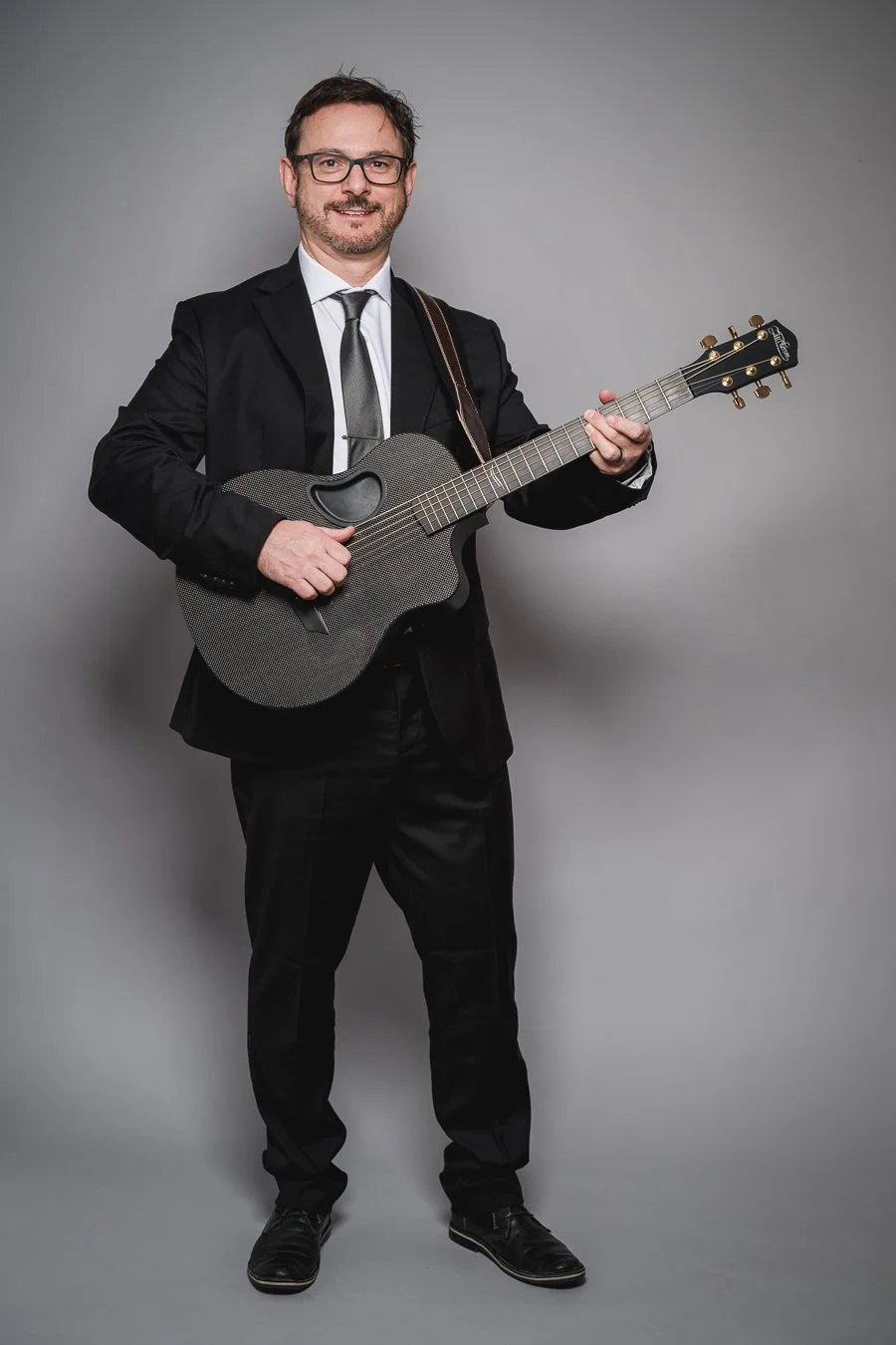 Man in a black suit and glasses playing a black acoustic guitar against a gray background.