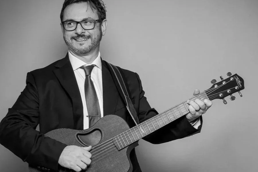 A man with glasses and a beard, dressed in a suit and tie, smiling while holding an acoustic guitar.