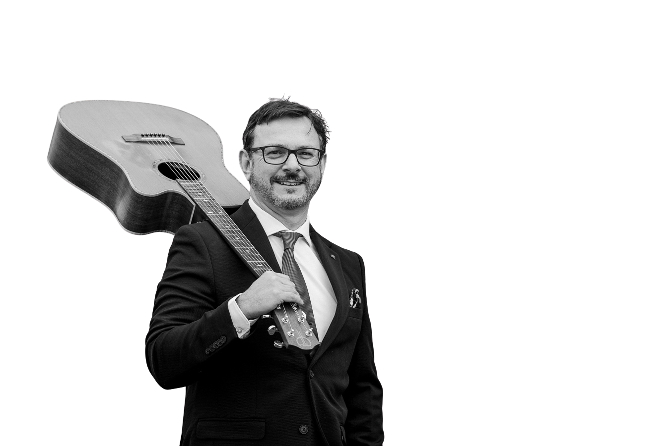 A man in a suit and glasses smiling while holding a guitar over his shoulder, with a plain black background.