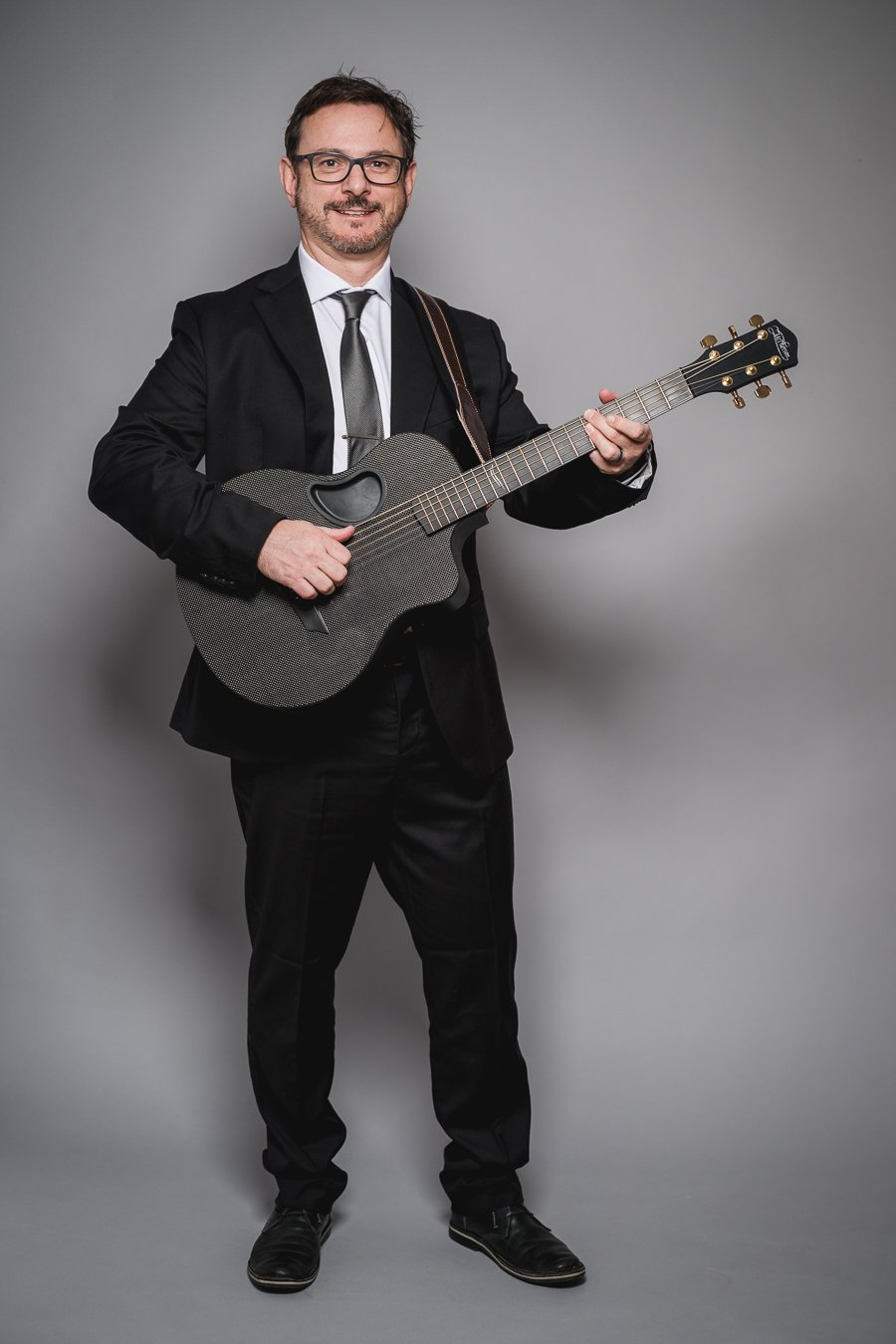 Man in a black suit and glasses playing a black acoustic guitar against a plain gray background.