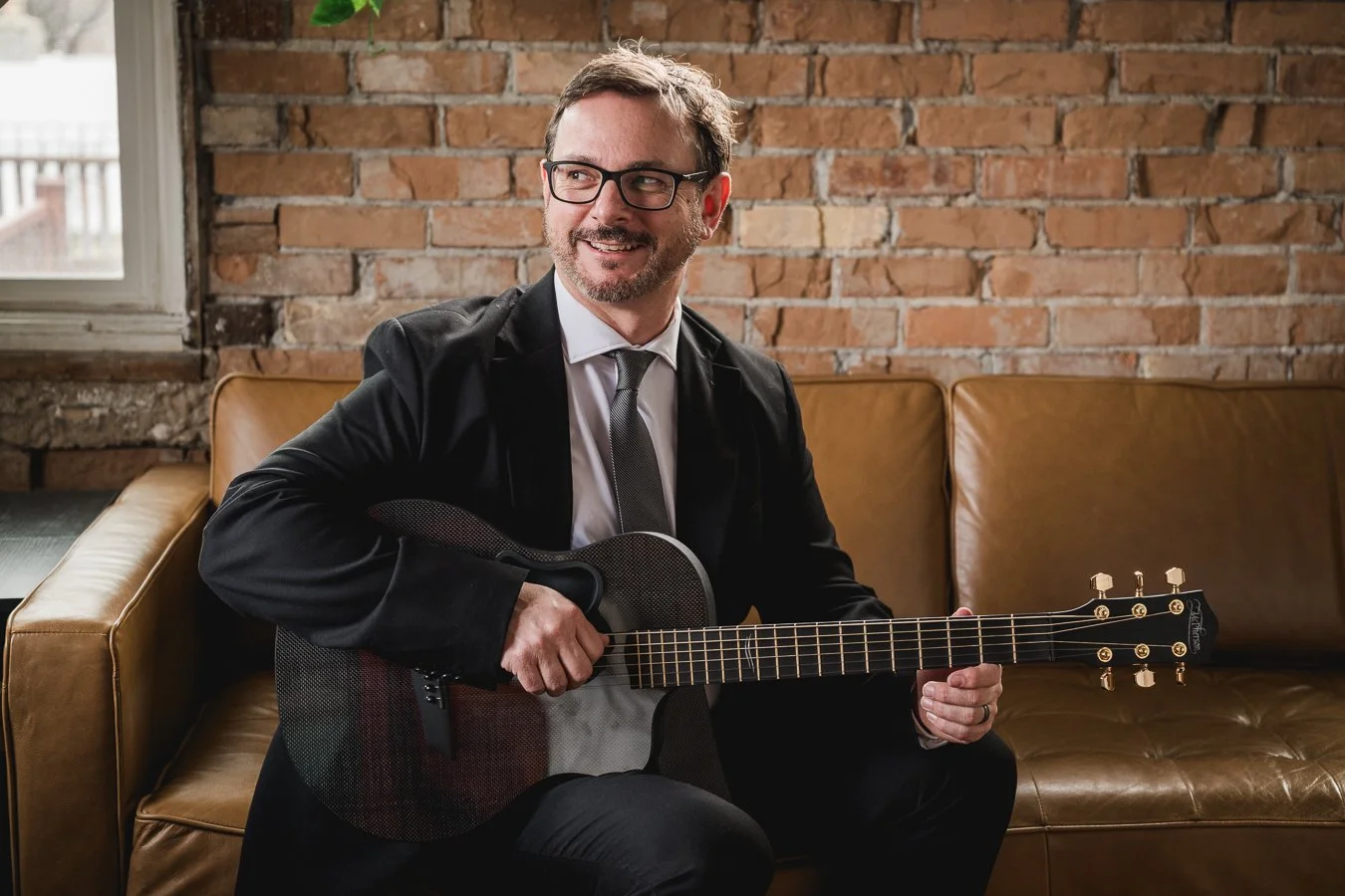 Man in a black blazer and glasses sitting on a leather sofa playing an acoustic guitar in a room with brick wall background.