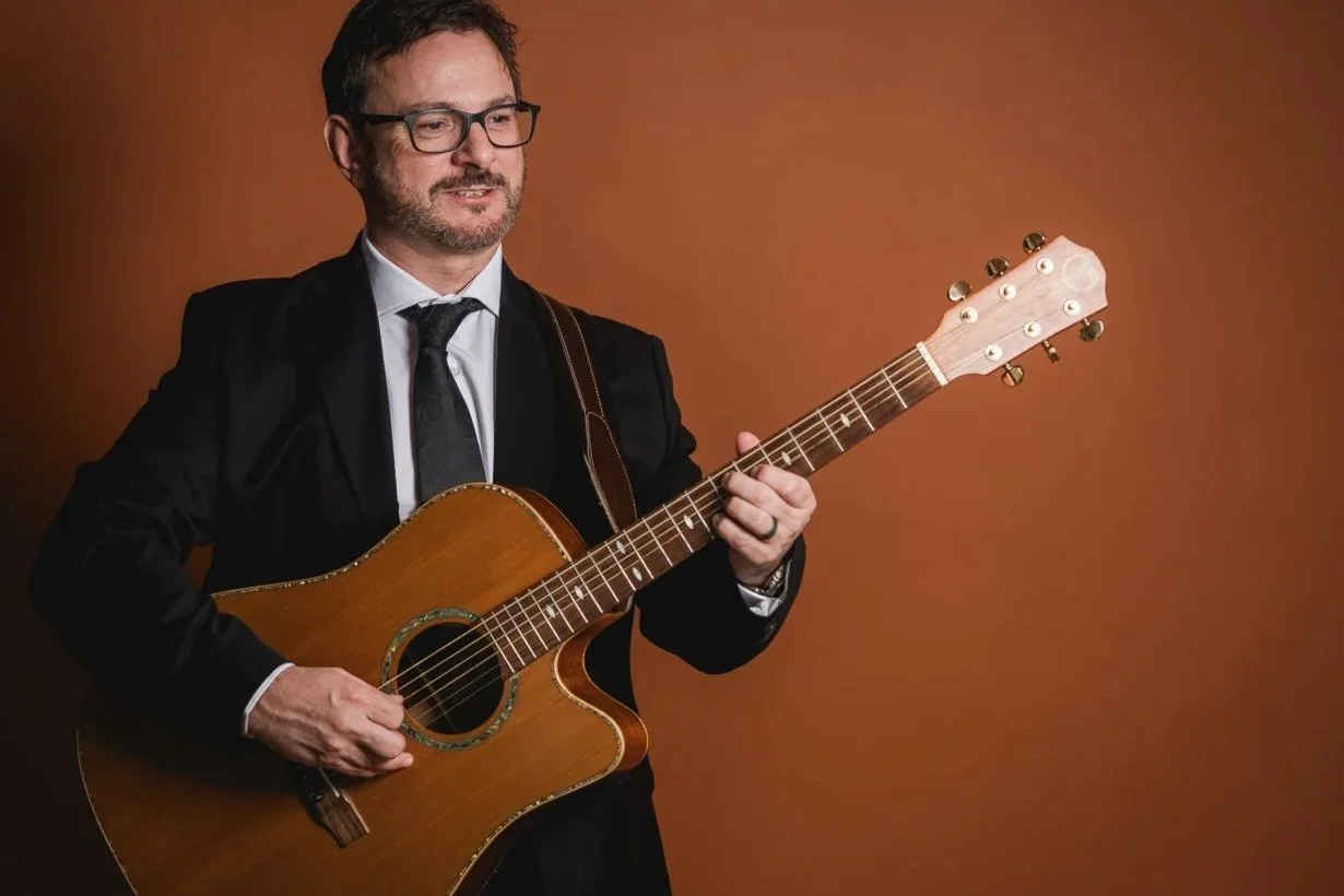 A man in a black suit and glasses playing an acoustic guitar against a brown background.