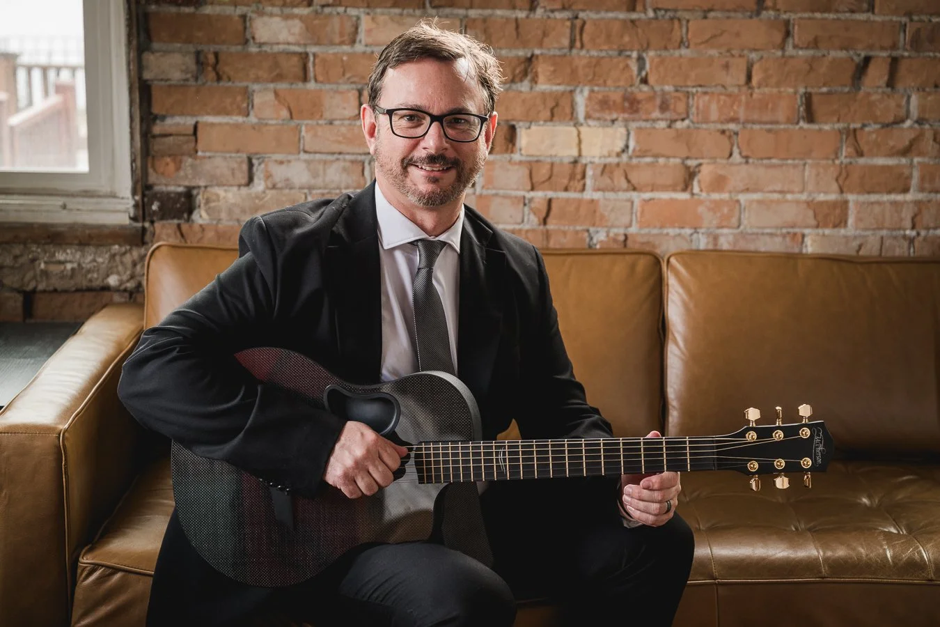 A man in a suit and tie, wearing glasses, sitting on a brown leather couch holding an acoustic guitar, with a brick wall background and a window to the side.