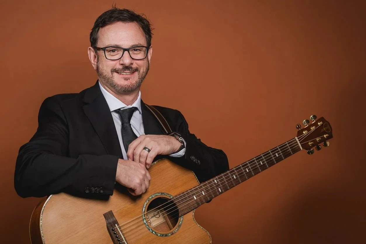 A smiling man with glasses and a beard in a black suit, white shirt, and black tie holding an acoustic guitar against a brown background.