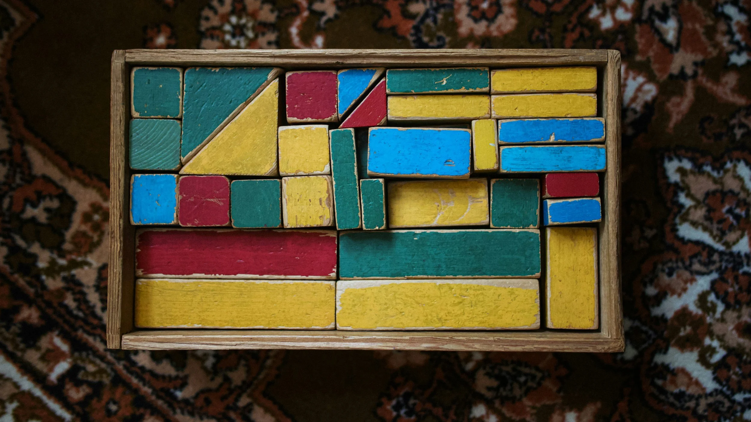 Top-down view of a wooden box filled with colorful wooden toy blocks arranged in various shapes and sizes, with a patterned rug underneath.