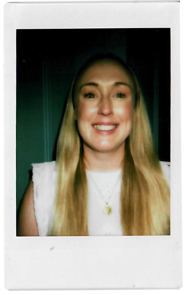 A woman with long blonde hair wearing a white top and a necklace with a round pendant, smiling at the camera in a selfie.