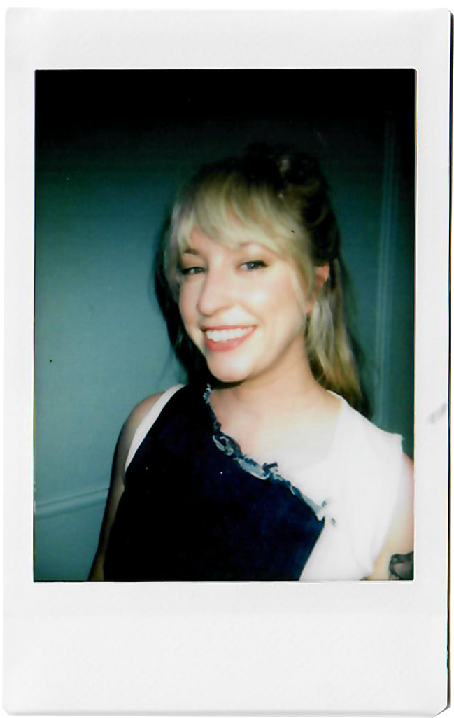A young woman with blonde hair, smiling, wearing a white and black sleeveless top, posing indoors.