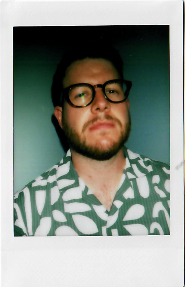 A man with dark hair, a beard, and glasses wearing a patterned shirt in front of a plain wall.