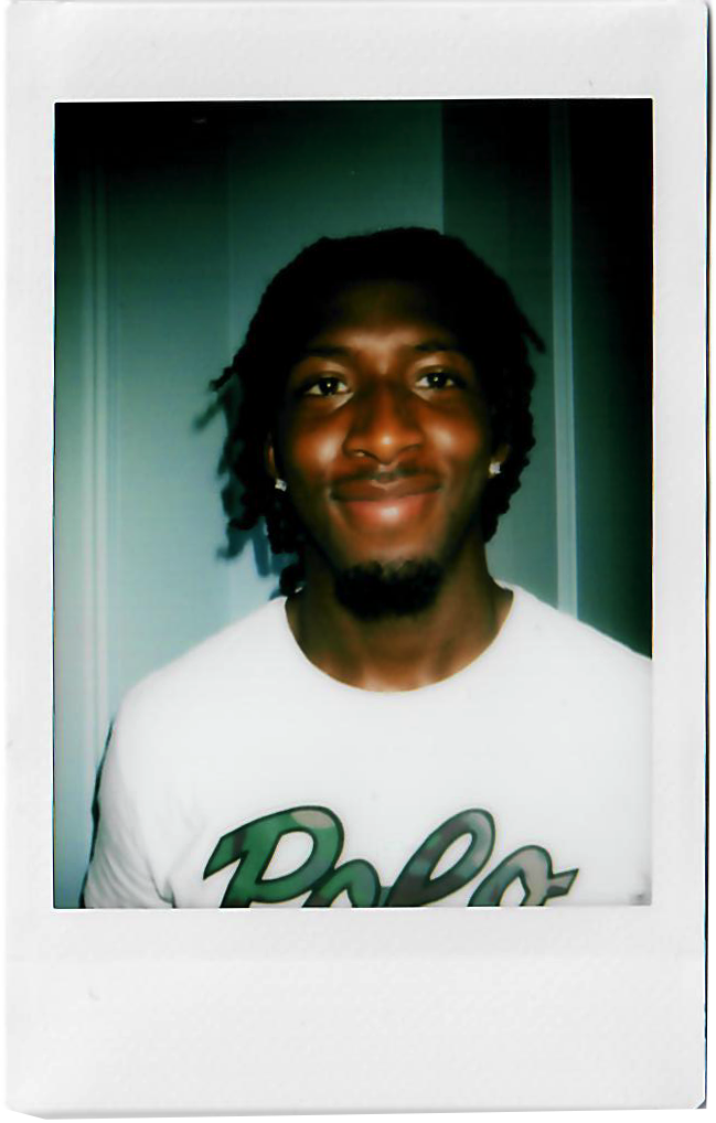 A young man with dark skin, dreadlocks, and a beard, wearing a white T-shirt with green lettering, smiling in front of a plain background.