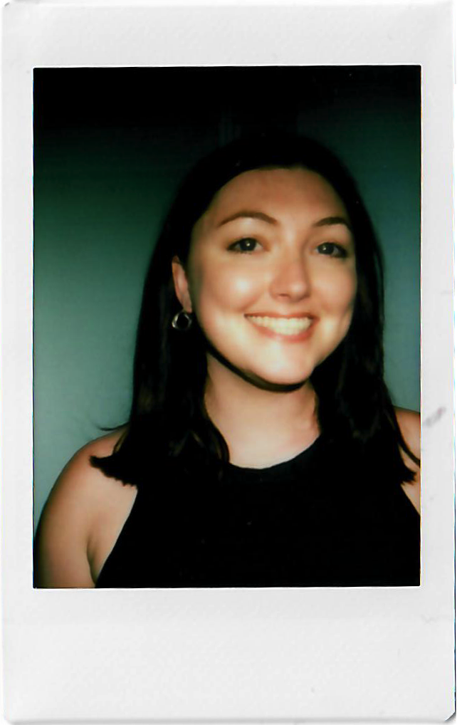 A young woman smiling with dark hair, earrings, wearing a black sleeveless top, and a black choker necklace, posing against a dark background.