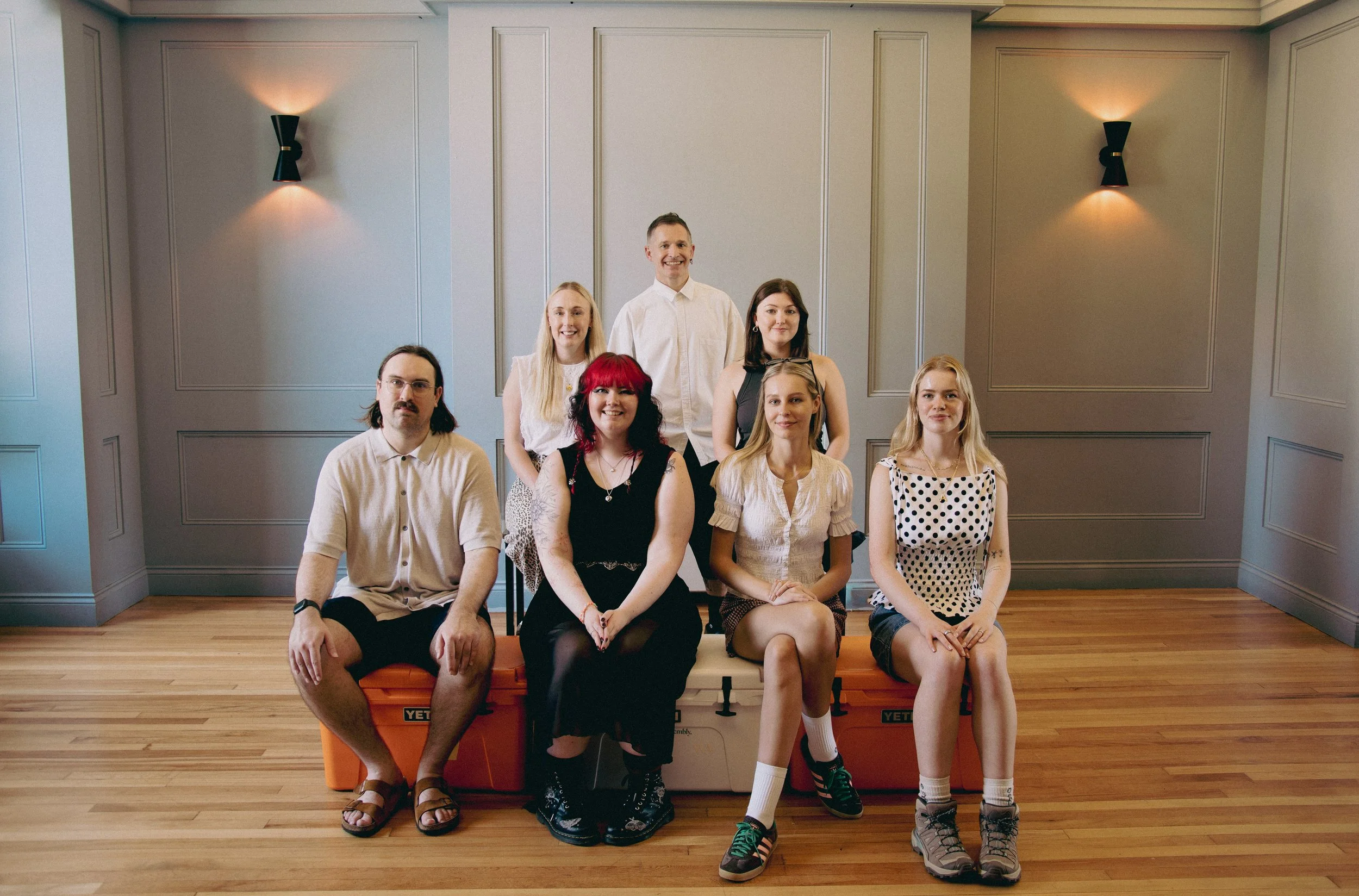 Group of seven young adults posing together in a room with gray walls and wooden flooring, sitting and standing around a large orange YETI cooler.