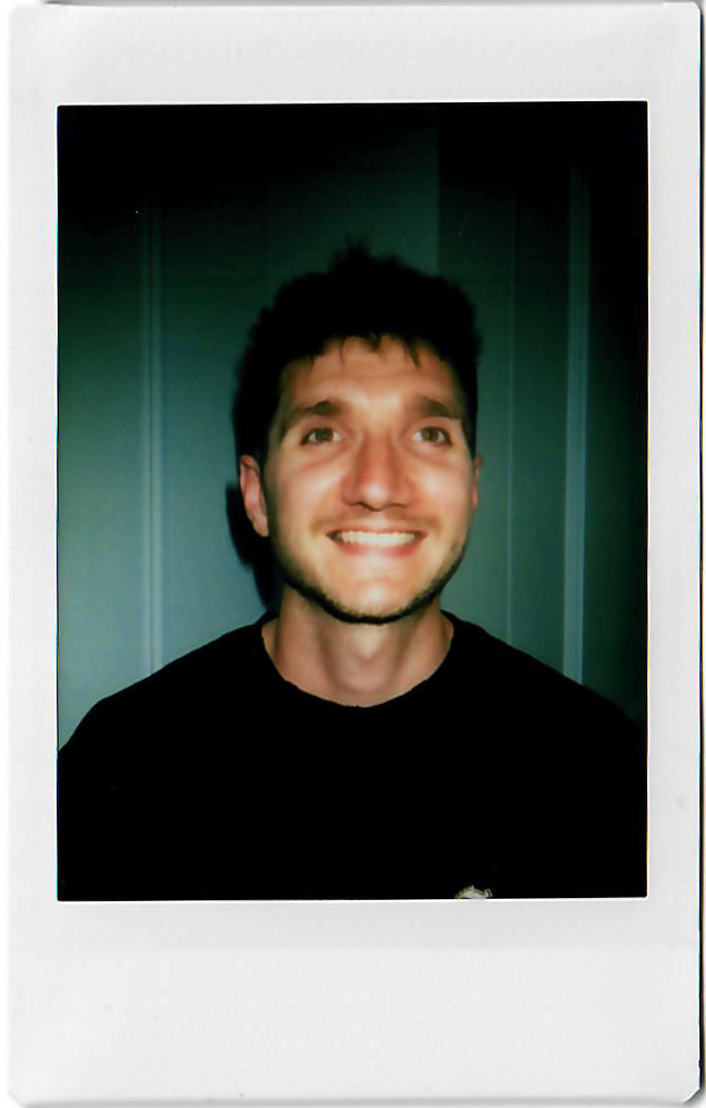 Young man with dark hair and a beard smiling, wearing a black shirt, standing in front of a dark wall with vertical panels.