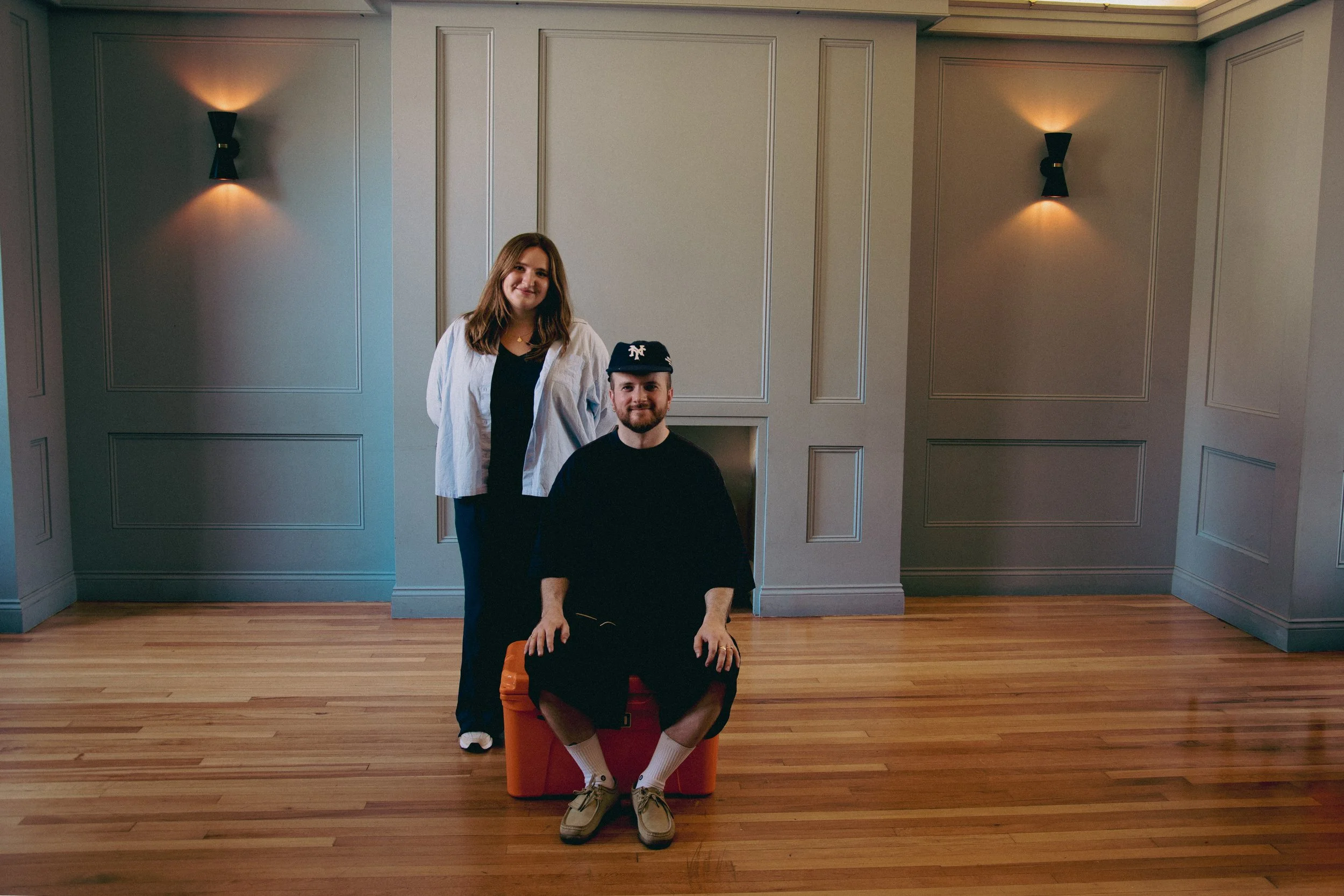 Two people, a woman and a man, posing in a room with wooden floors and gray paneled walls. The woman is standing behind the man, who is sitting on an orange stool, with a smile. The man is wearing a black shirt, shorts, sneakers, and a cap, while the woman is wearing a white shirt over a black top.