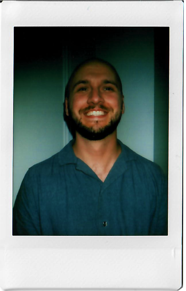 Photograph of a smiling man with a beard, wearing a blue shirt, standing indoors against a plain wall.