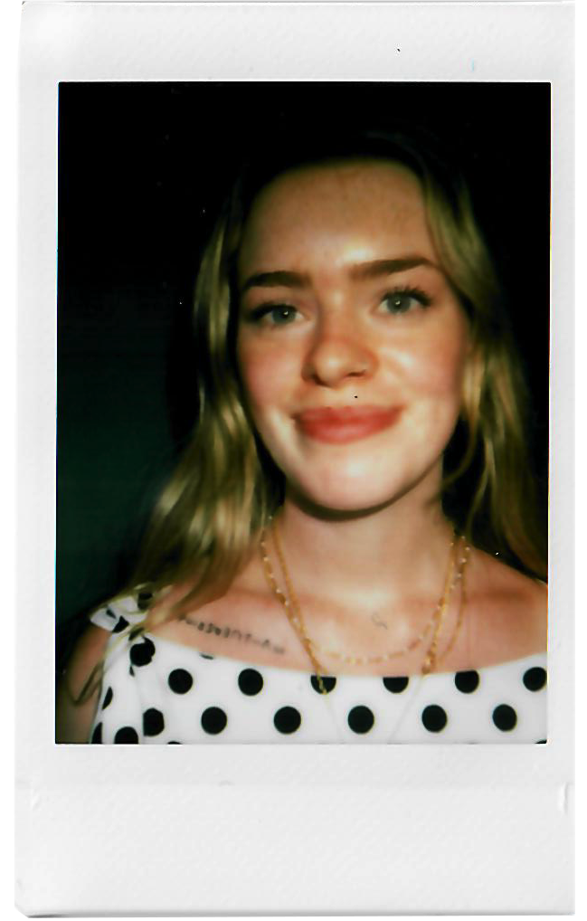 A young woman with blonde hair, smiling, wearing a black and white polka dot sleeveless top, gold necklaces, and a small nose piercing.