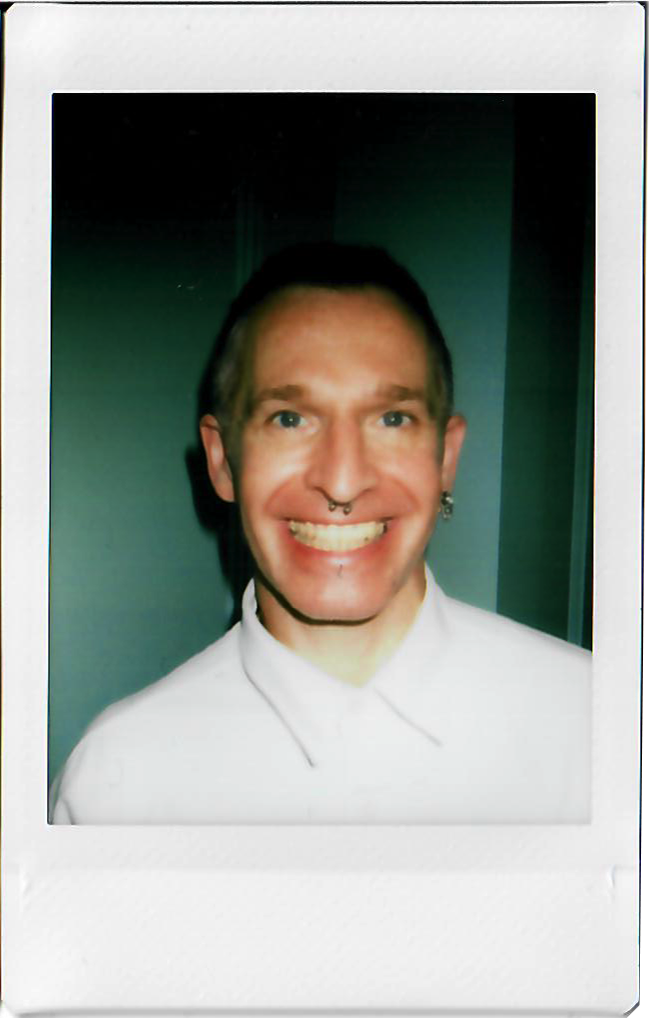 A smiling man with blue eyes, pale skin, and piercings, wearing a white collared shirt, standing against a dark background