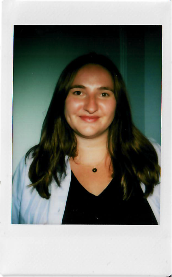 A young woman with long brown hair smiling, wearing a black top, a white shirt draped over her shoulders, and a necklace with a small pendant.