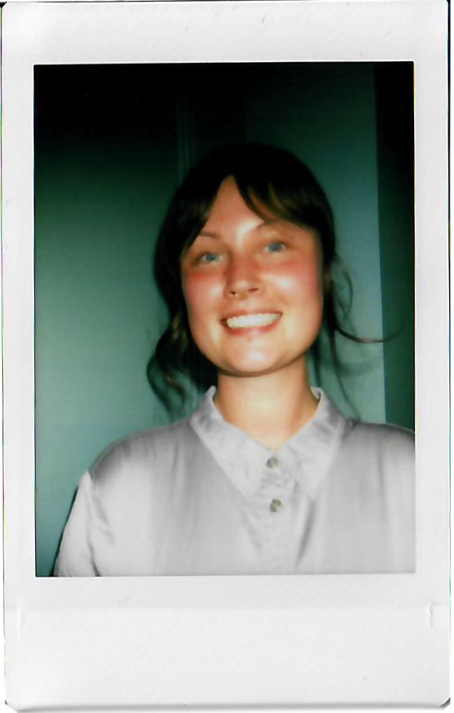 A woman with shoulder-length dark hair, light skin, and blue eyes, smiling and wearing a light-colored collared shirt, standing in front of a greenish background.