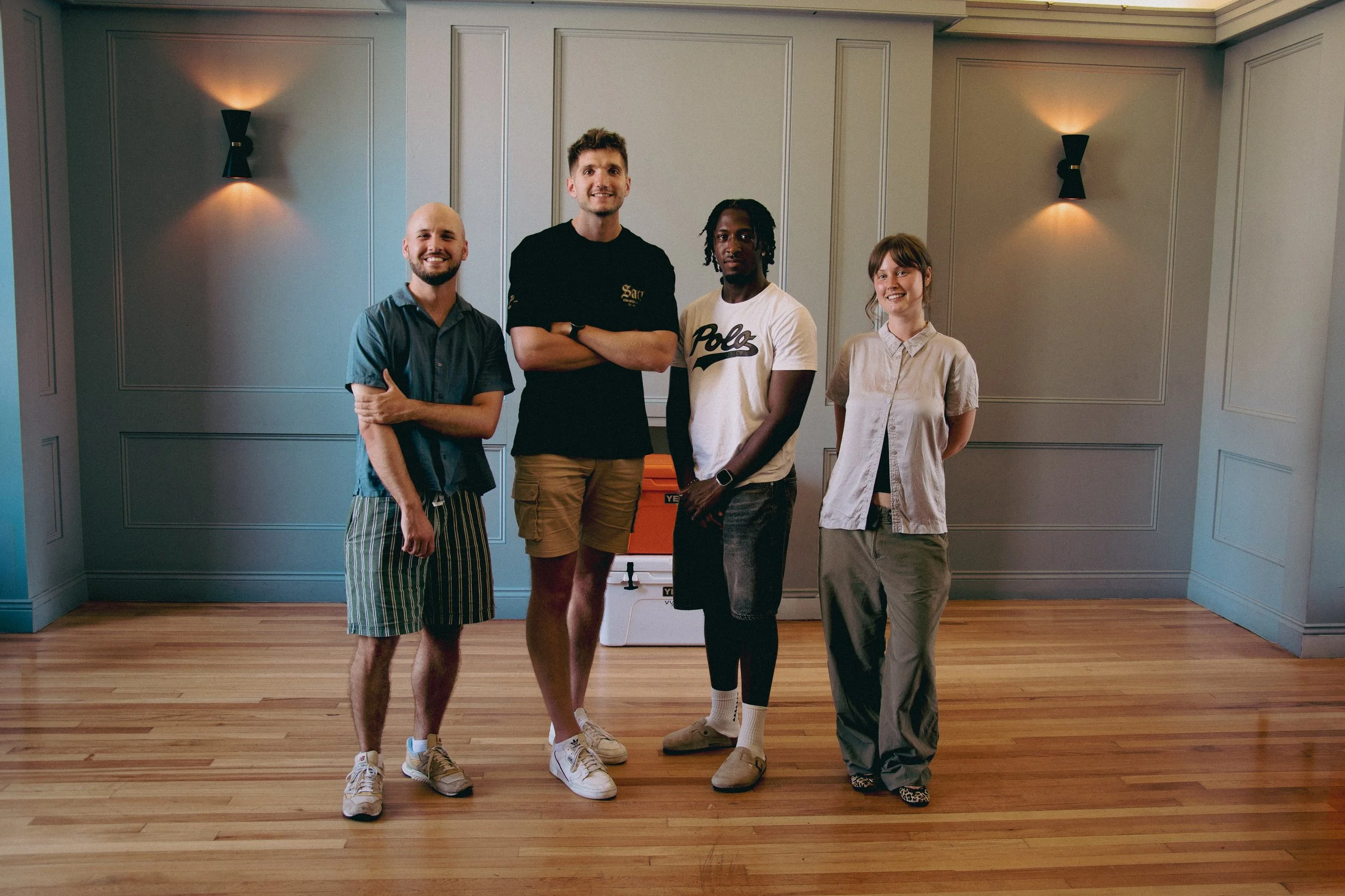 Four young adults standing in a room with wooden floors and decorated gray walls, smiling at the camera.