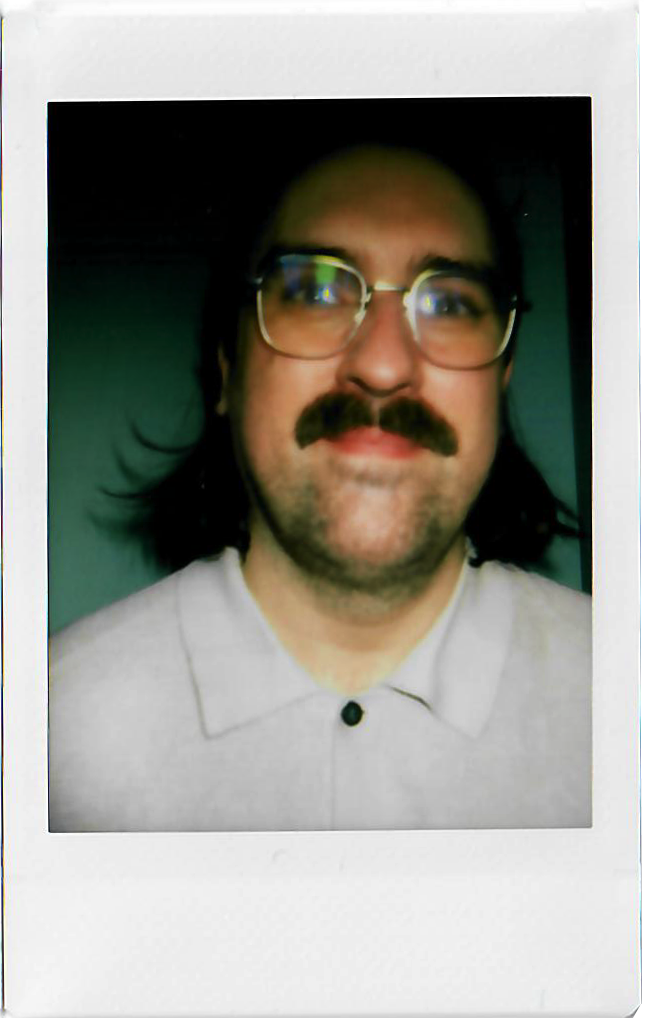A man with glasses and a mustache taking a selfie, wearing a light-colored shirt.