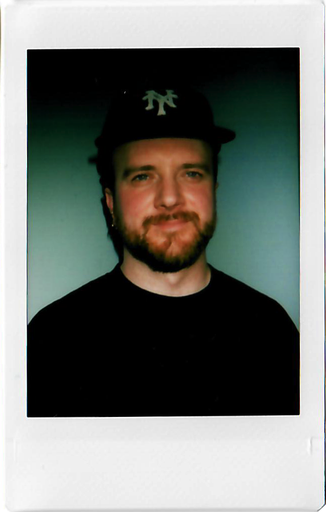 A young man with a beard and mustache wearing a black New York Yankees baseball cap and black shirt, smiling slightly against a neutral background.