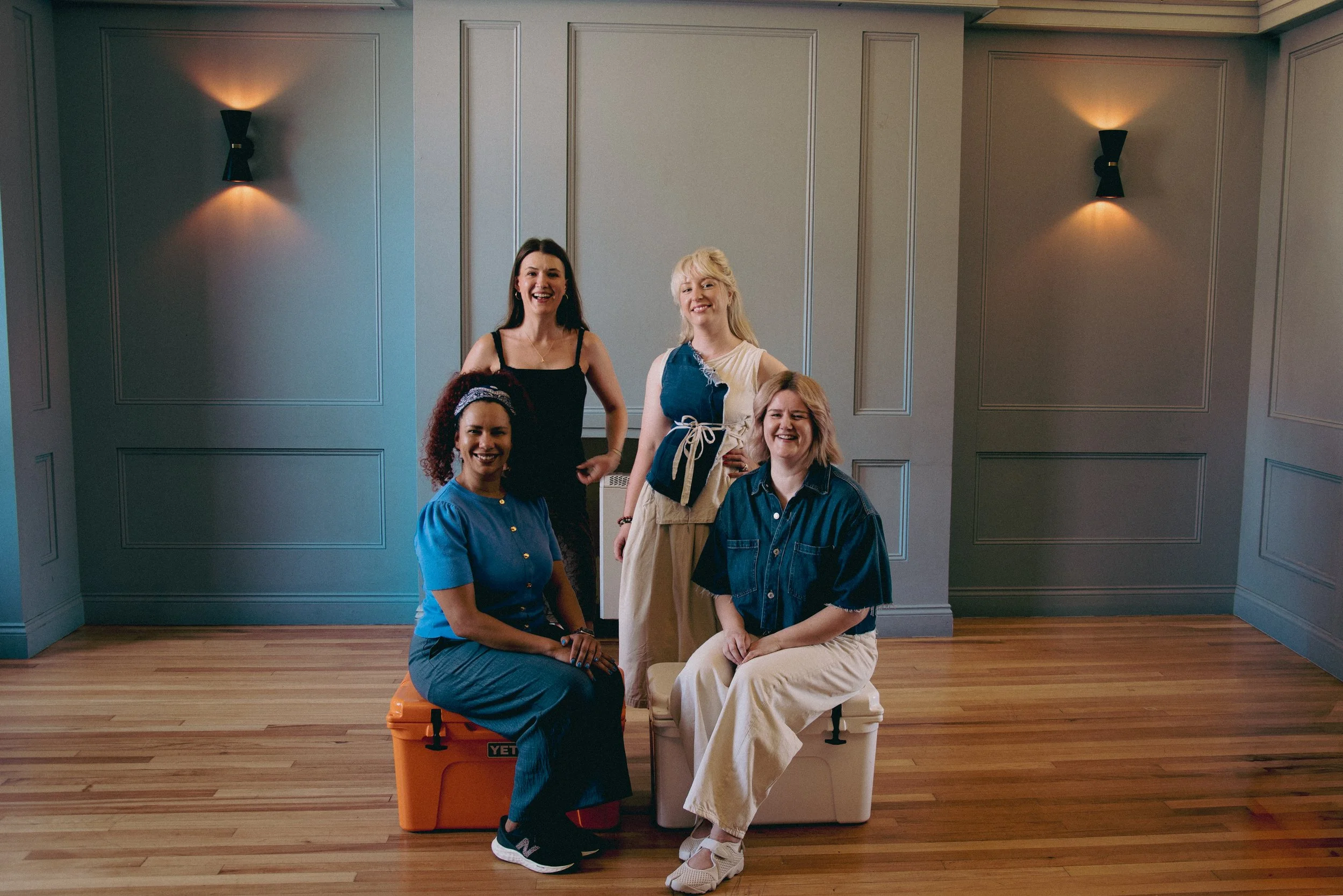 Five women posing together indoors against a gray paneled wall, some seated on a plastic storage container, smiling at the camera.