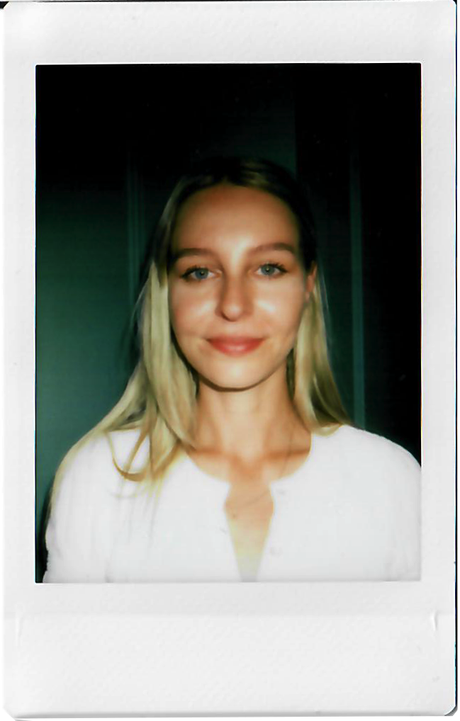 A young woman with blonde hair and blue eyes, smiling and wearing a white top, taking a portrait photo indoors.