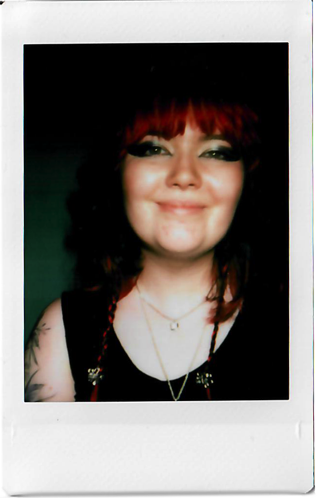 Young woman with red hair and tattoos smiling in a black top, wearing layered necklaces.