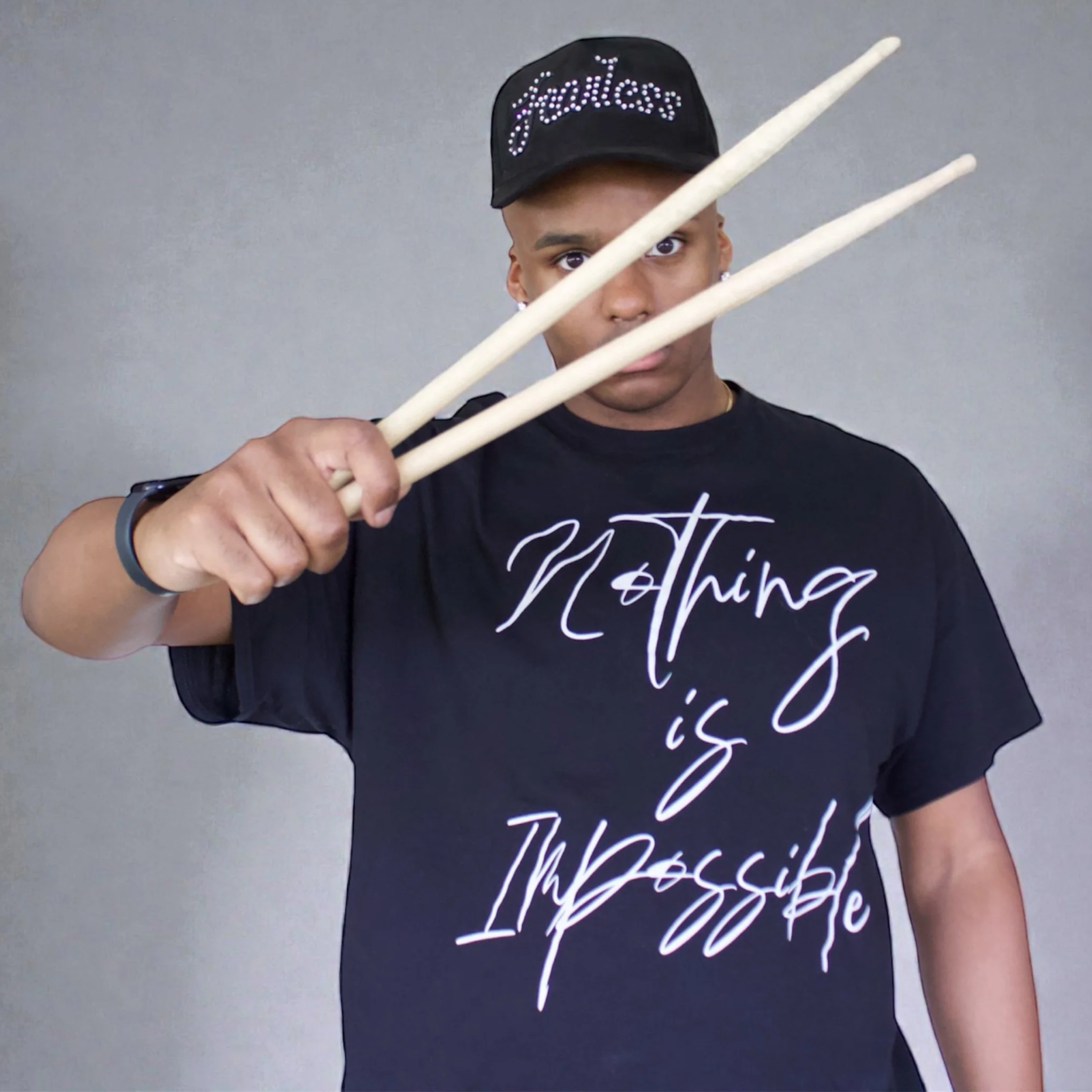 Portrait of Jordan in a black shirt and black hat, holding two drumsticks in his hand.
