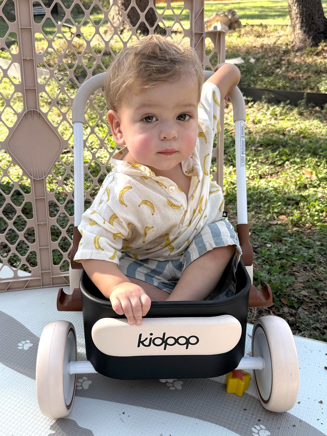 A young child with light brown hair and fair skin sitting in a KidGap stroller. The child is wearing a white shirt with a banana print and plaid shorts, sitting outdoors on a white surface with paw prints, surrounded by greenery and sunlight.