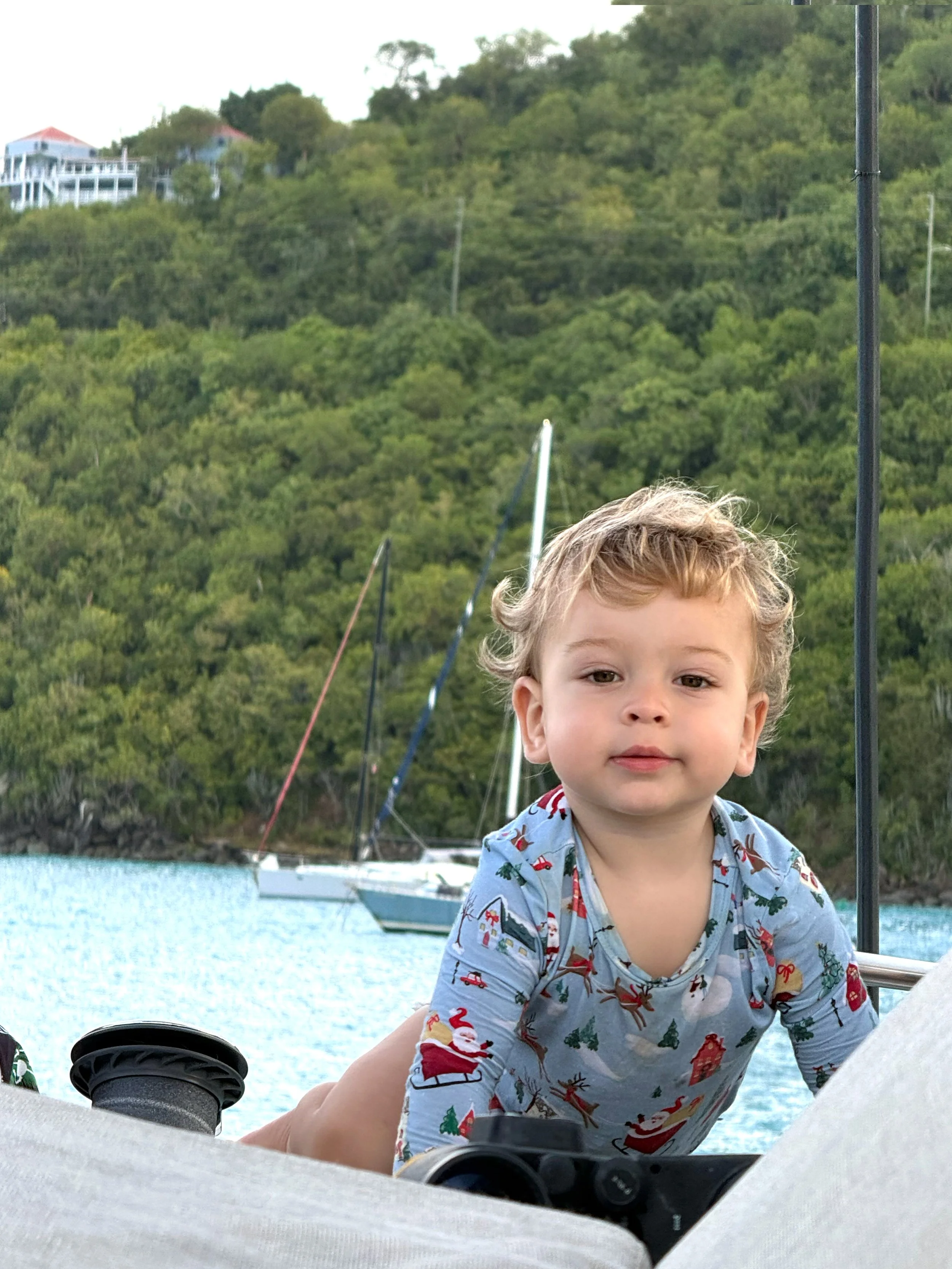 Young child with curly hair wearing a Christmas-themed shirt, sitting on a boat with sailboats and green hillside in the background.