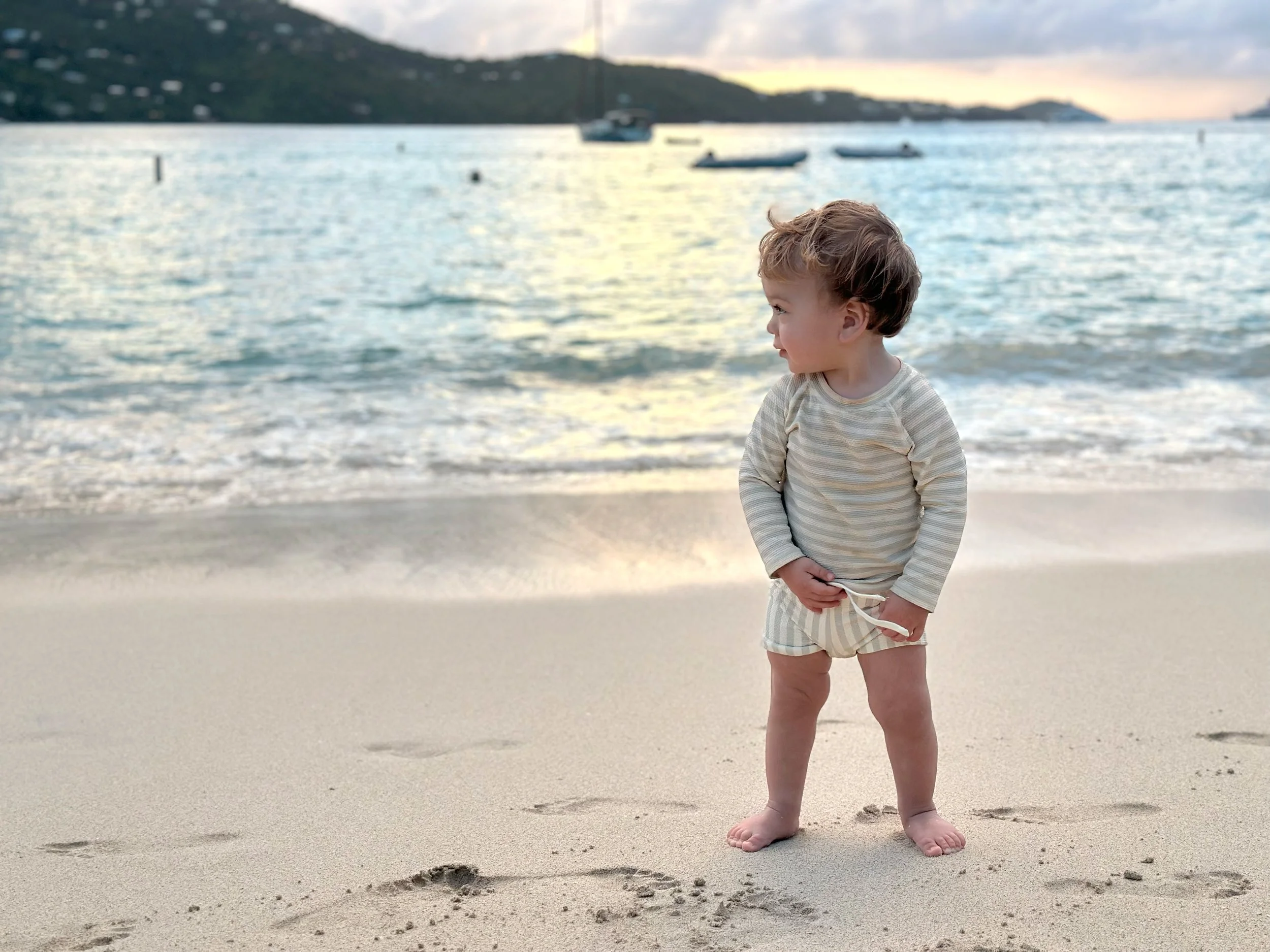 A young boy standing barefoot on the sandy beach near the shoreline, holding sunglasses with a harbor and boats in the background during sunset.