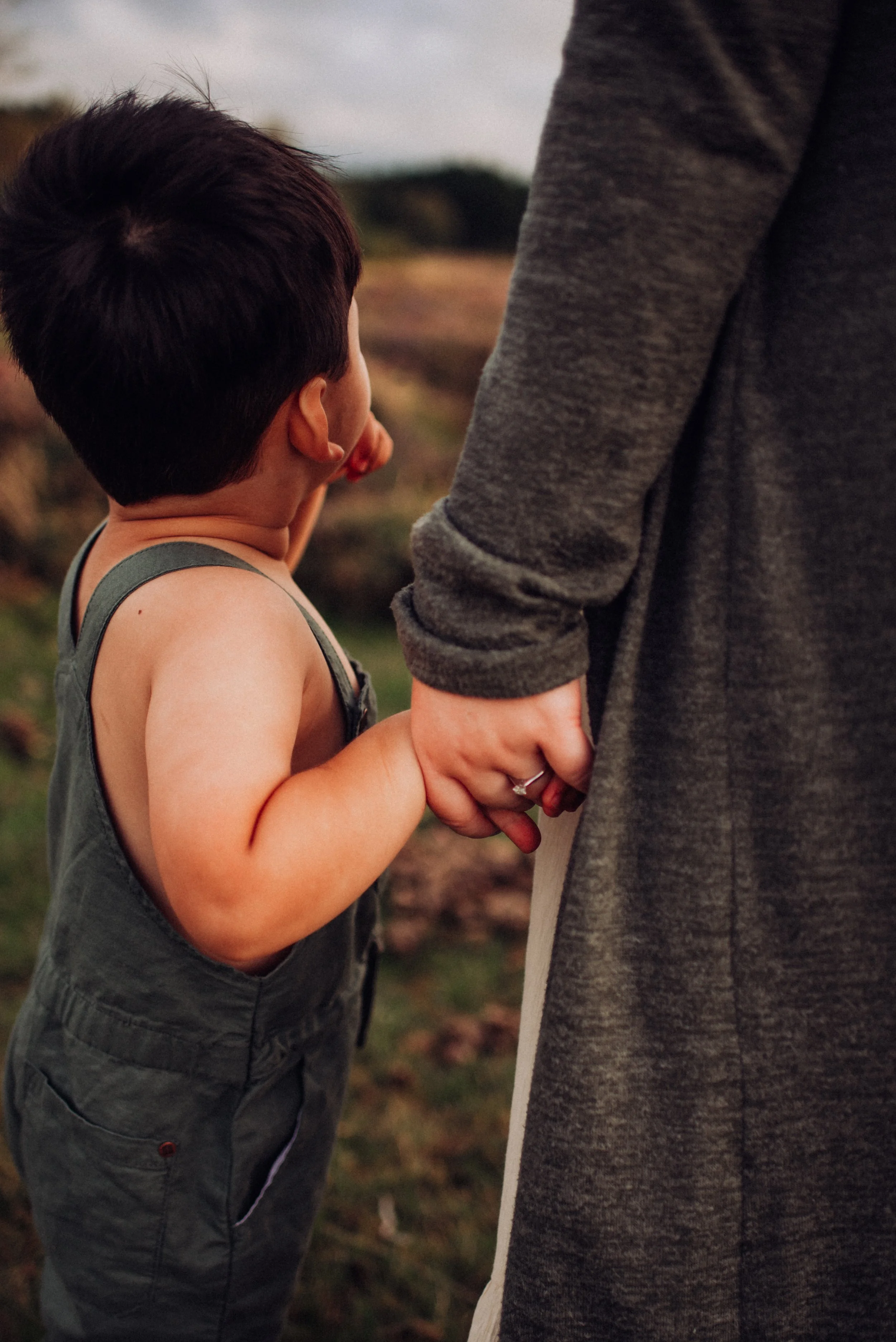 A young boy holding hands with an adult, possibly his mother or father, outdoors in a natural setting during daytime, with a blurred background of grass and trees.