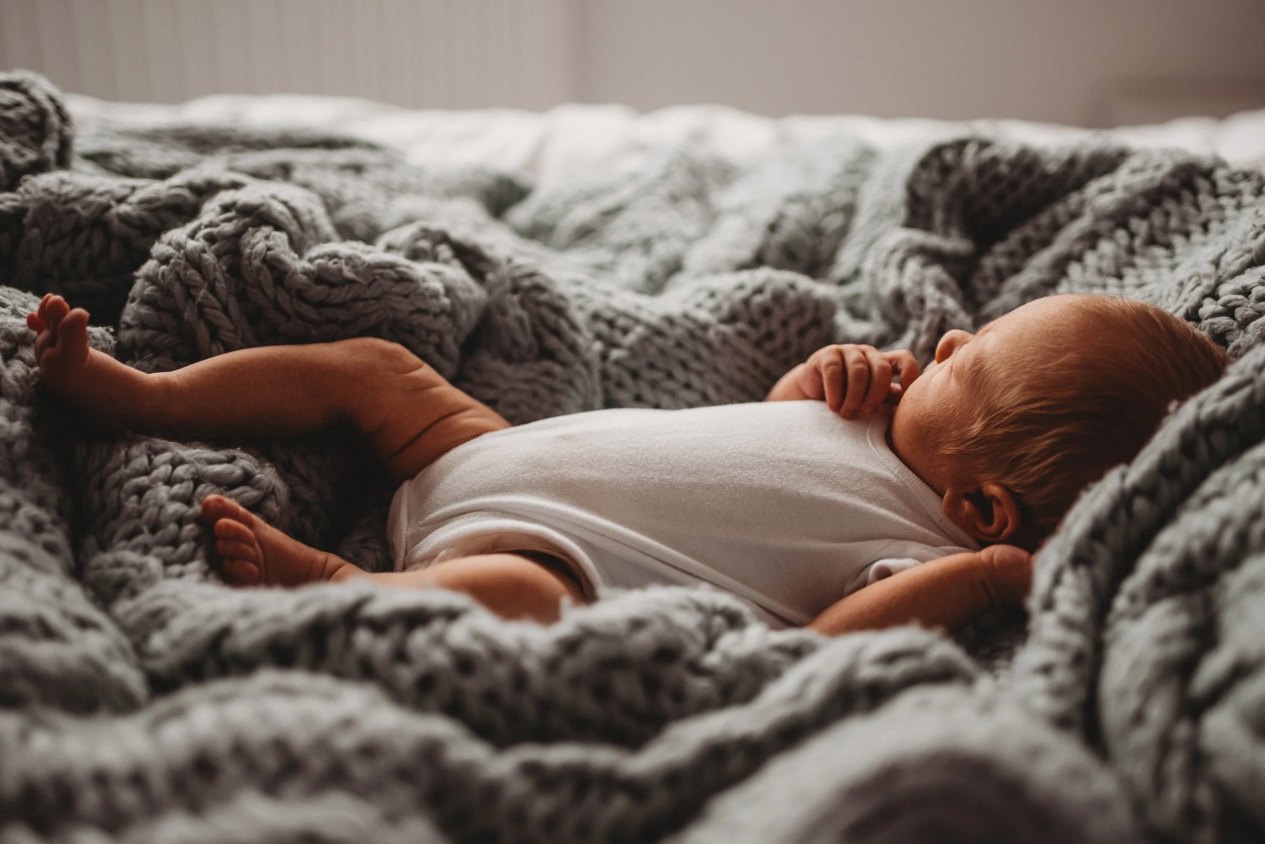 Lifestyle newborn photography of a  sleeping baby lying on a bed surrounded by large, soft, chunky gray knitted blankets.