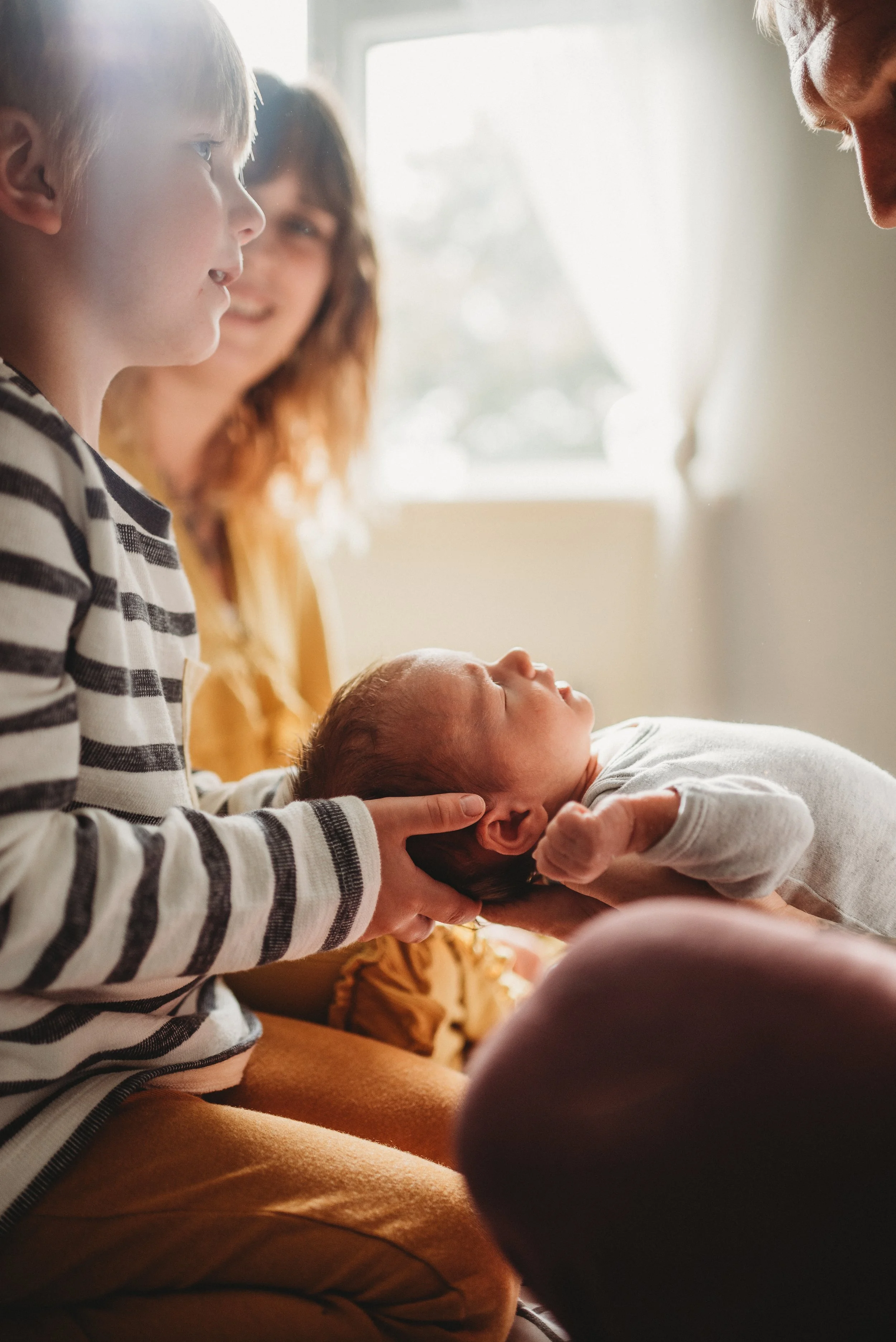 A woman and a young boy hold a newborn baby, cradling its head and shoulders, in a warmly lit room with a window in the background.