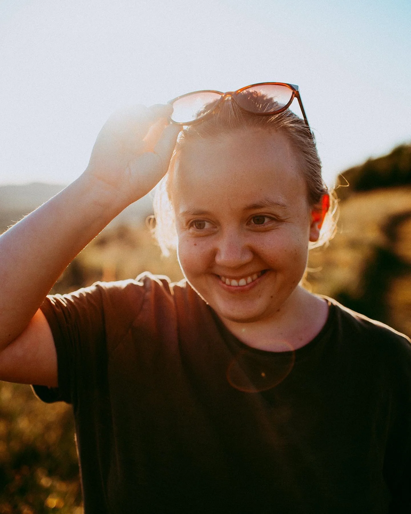 A smiling young woman holding sunglasses on her head outdoors during sunset.