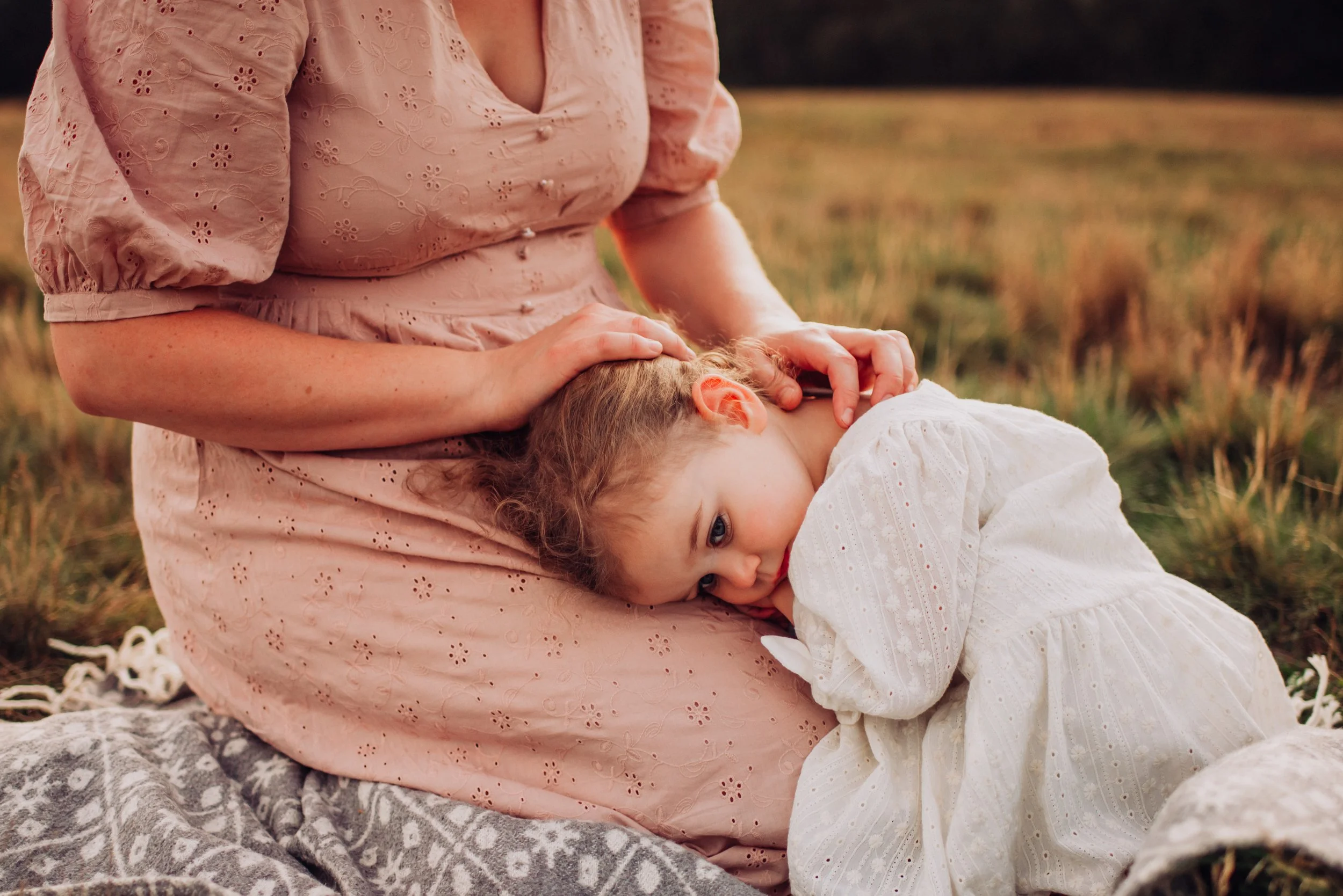 A woman in a pink dress sitting on a blanket with a young girl in a white dress, resting her head on the woman's lap in an outdoor grassy area.