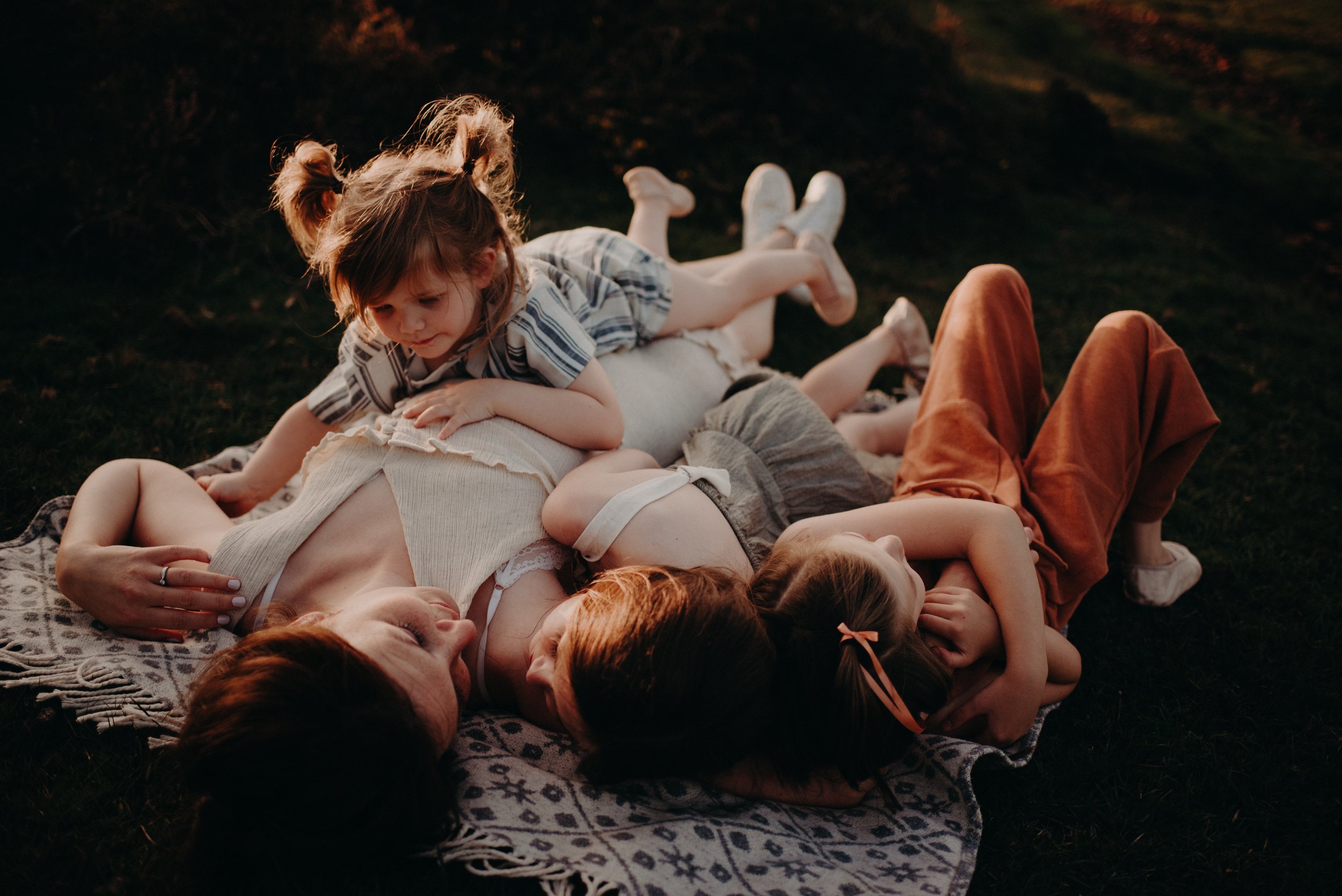 A mother lying on her back on a blanket on the grass, surrounded by three young children, as they play and cuddle together outdoors during sunset.