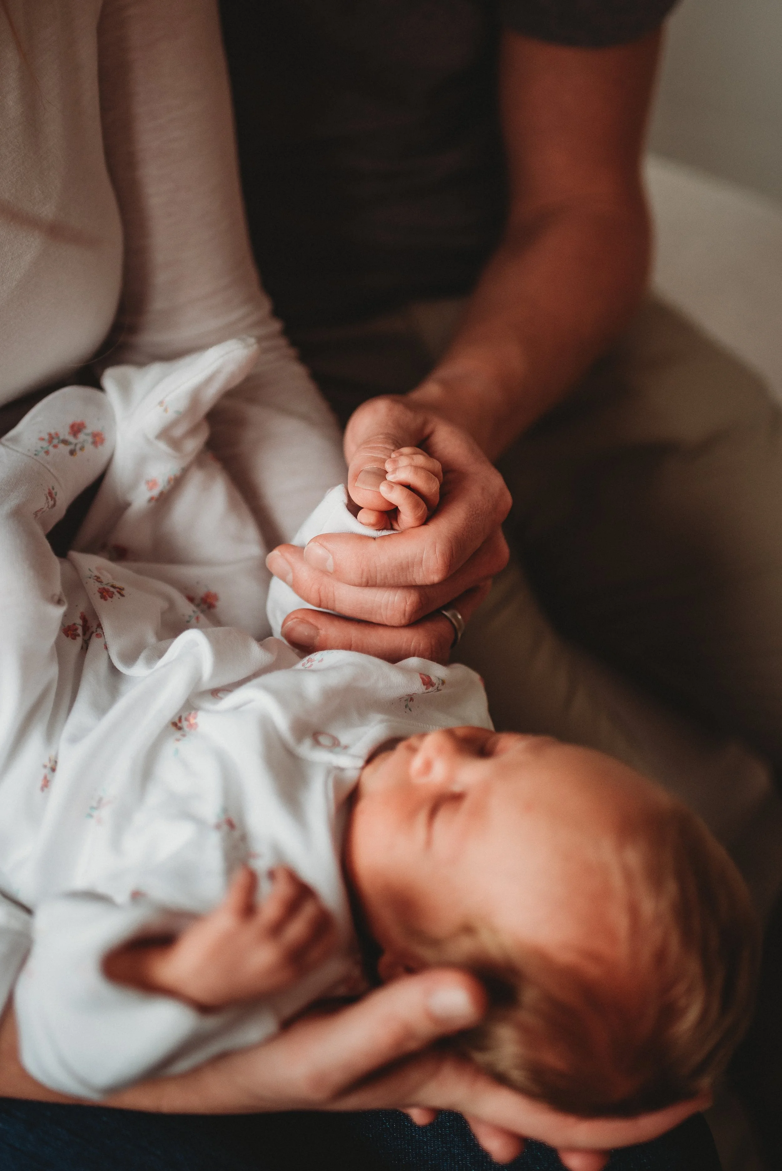 Lifestyle newborn photography of a person holding a baby's hand gently while the baby is lying down, dressed in a white outfit with floral patterns, with the baby's face resting peacefully.