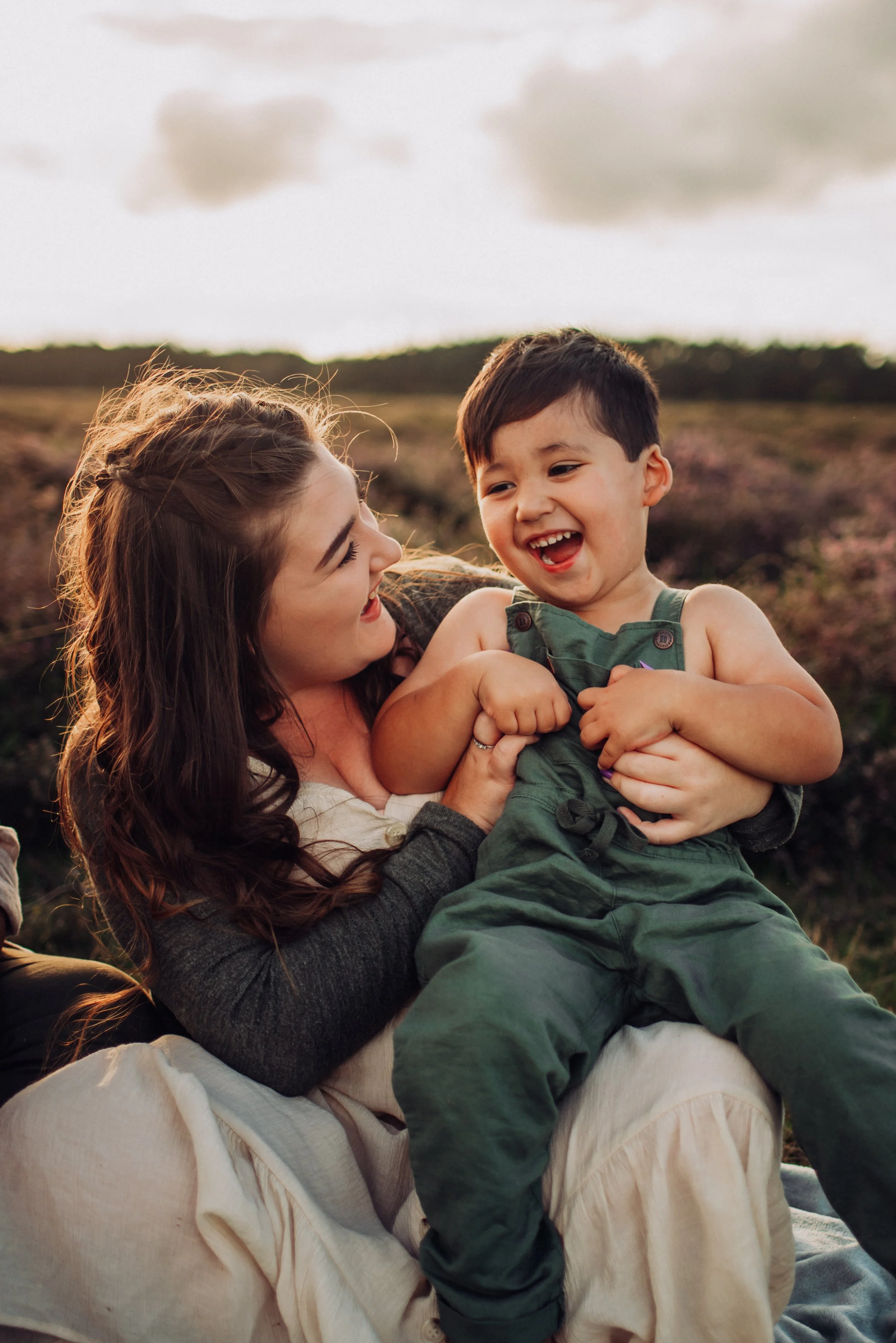 A woman and young boy are playing outdoors, smiling and laughing together in a field during sunset.