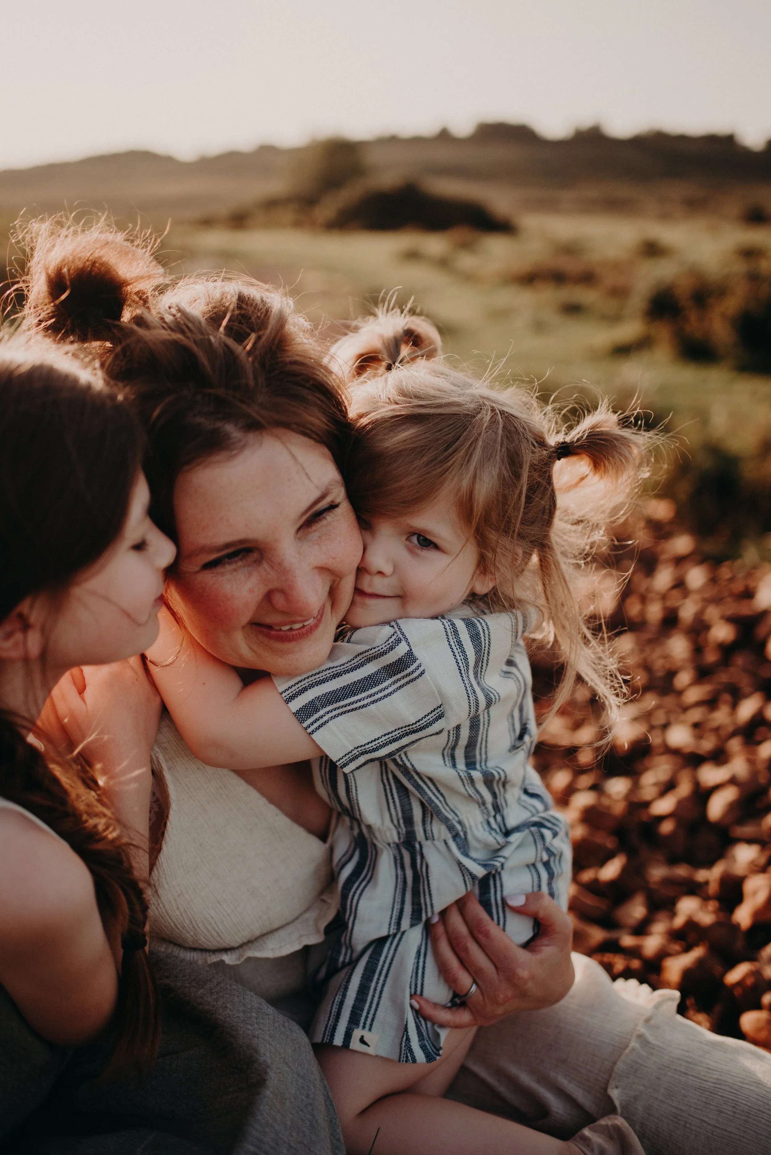 A woman with red hair embracing two young girls outdoors in a rocky landscape at sunset.