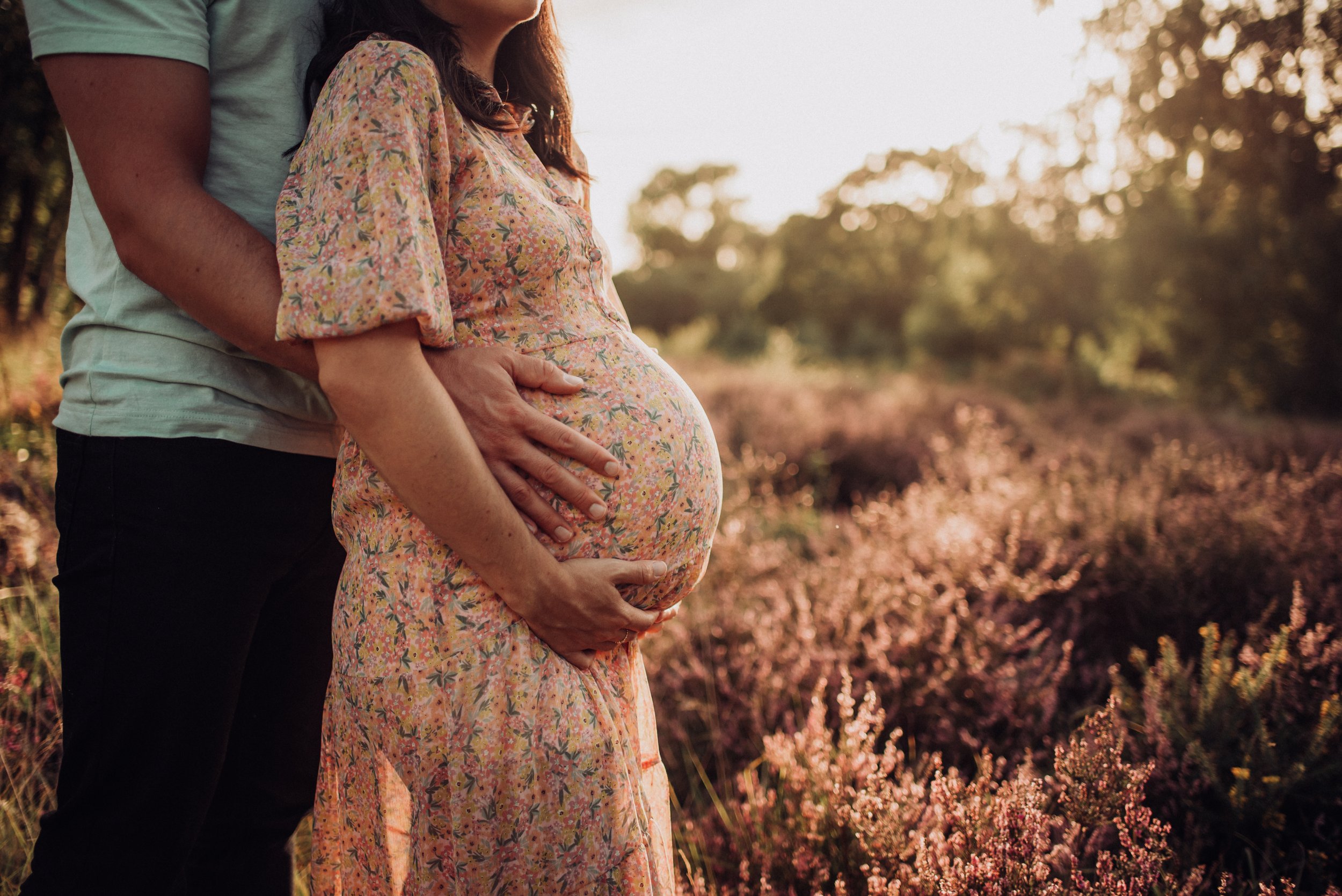 A pregnant woman wearing a floral dress standing outdoors with a man behind her, both with arms around her belly, outside in a natural setting during sunset.