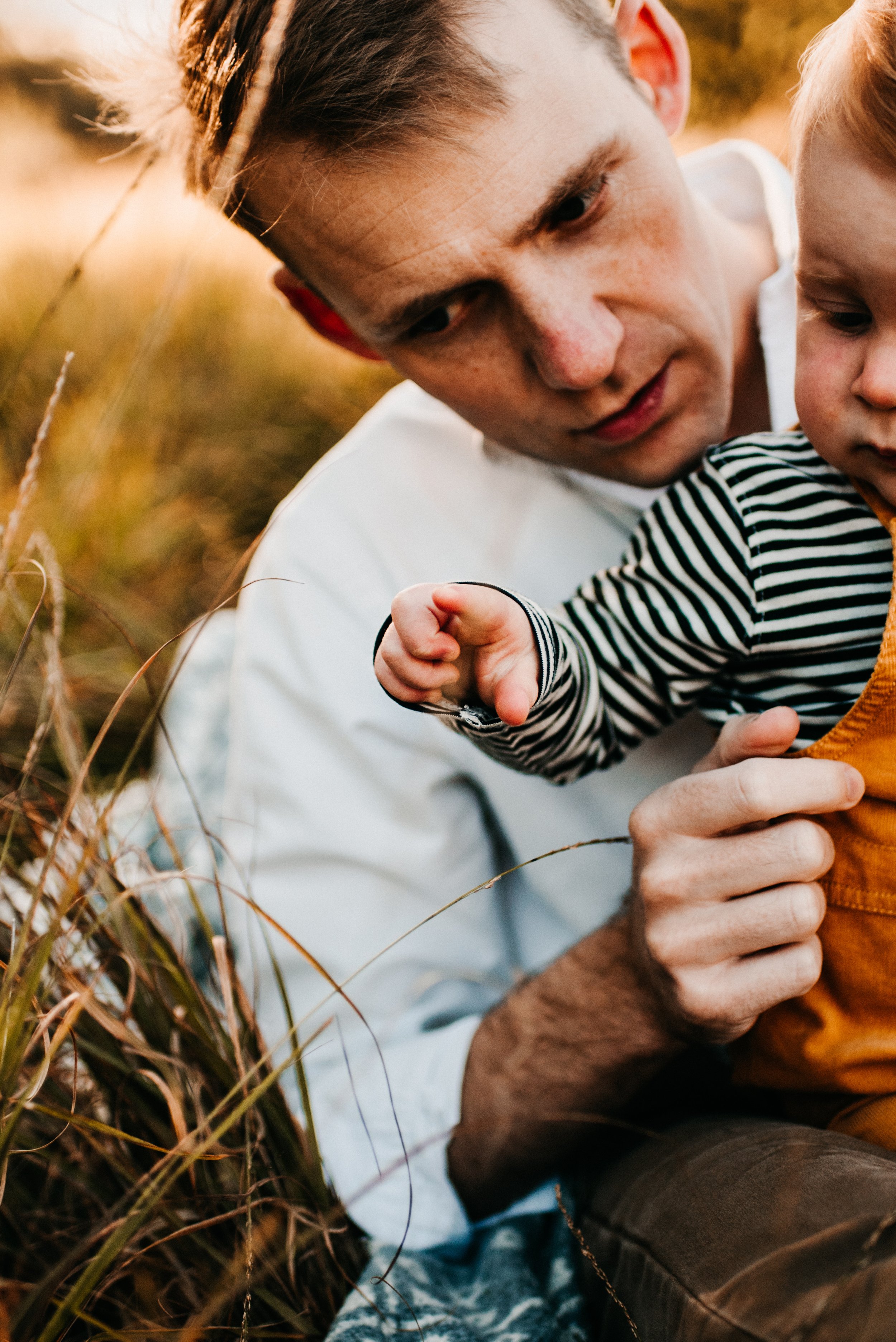 A young man and a small child sitting in tall grass outdoors, with the man showing the child something while they look at it closely.