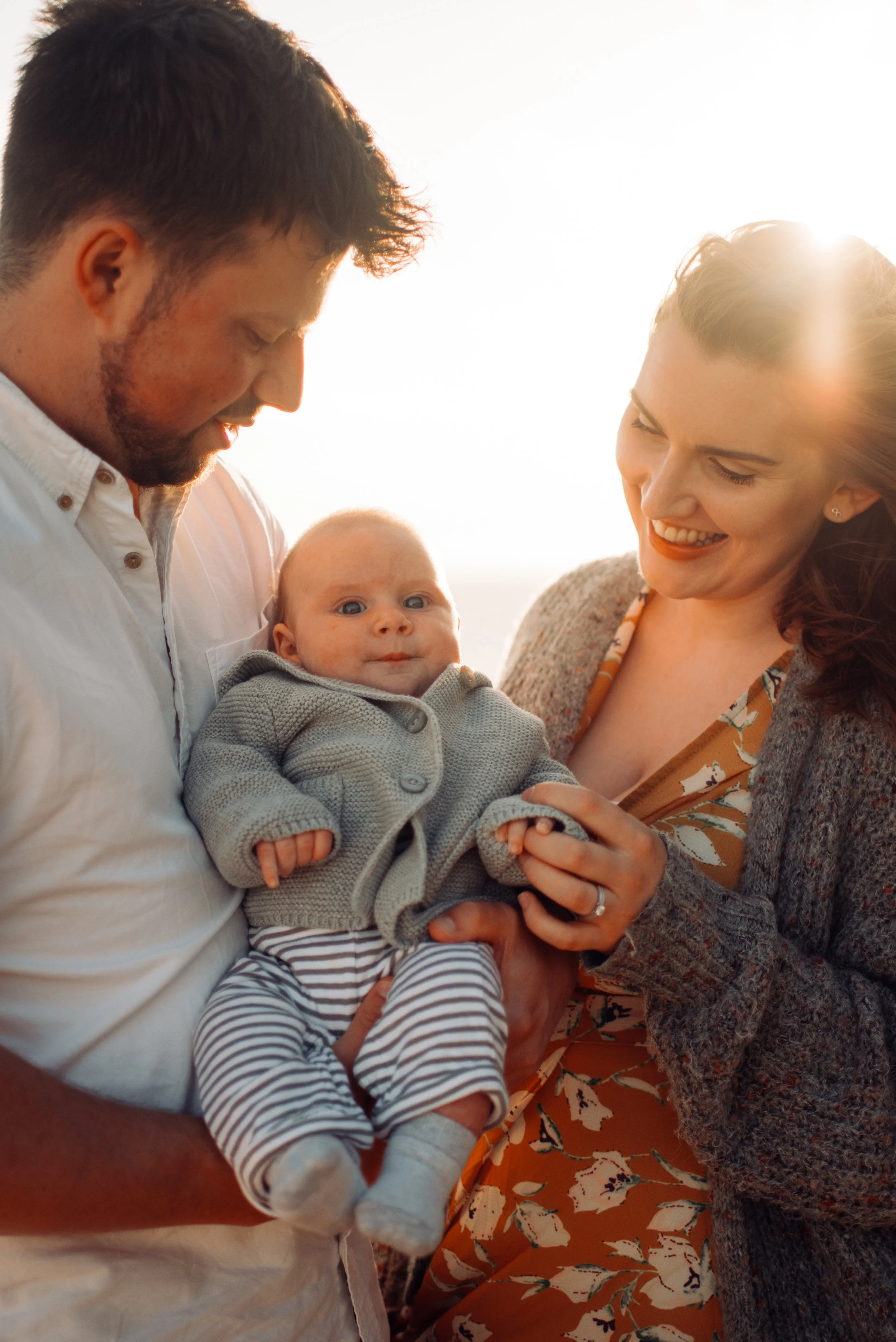 A happy family with a man, woman, and baby looking at the camera outdoors during sunset.
