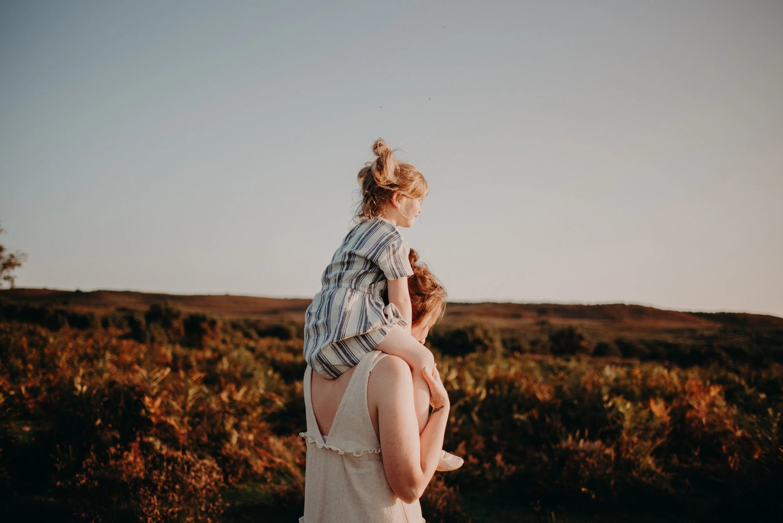 A woman carrying a young girl on her shoulders outdoors during sunset, with rolling hills and shrubbery in the background.