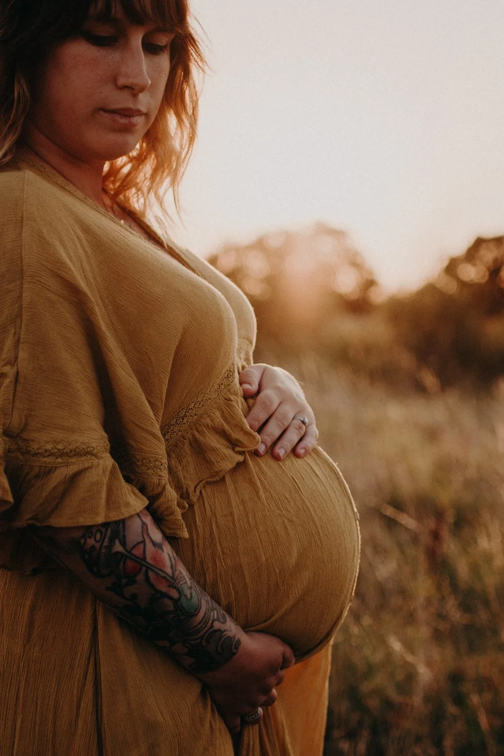 A pregnant woman in a mustard yellow dress stands outdoors during golden hour, cradling her belly with her right hand, her left hand resting beneath it. She has wavy, shoulder-length reddish hair and colorful tattoos on her left forearm. The background features blurred trees and earthy terrain.