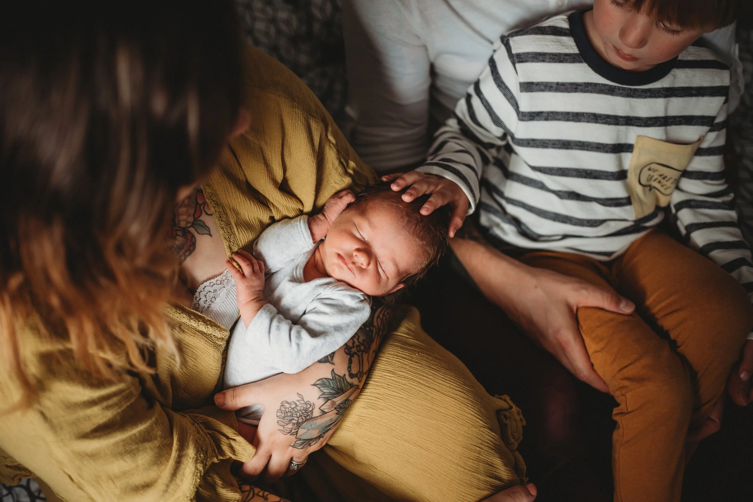 A newborn baby being held by an adult with tattoos, surrounded by two children. The baby is wearing a gray outfit and the adults and children are sitting close together.