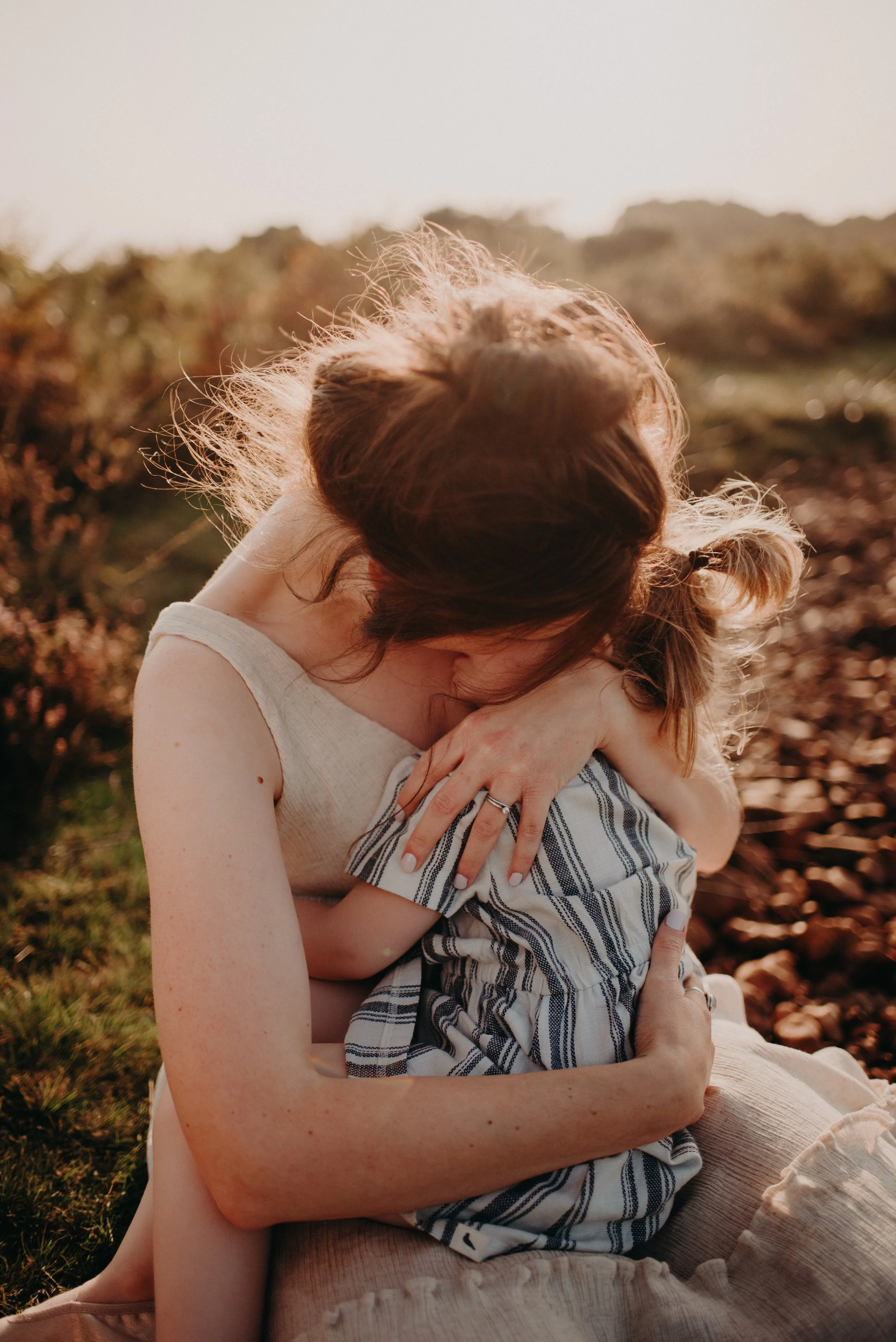 A woman with red hair and a young girl hugging outdoors during sunset.